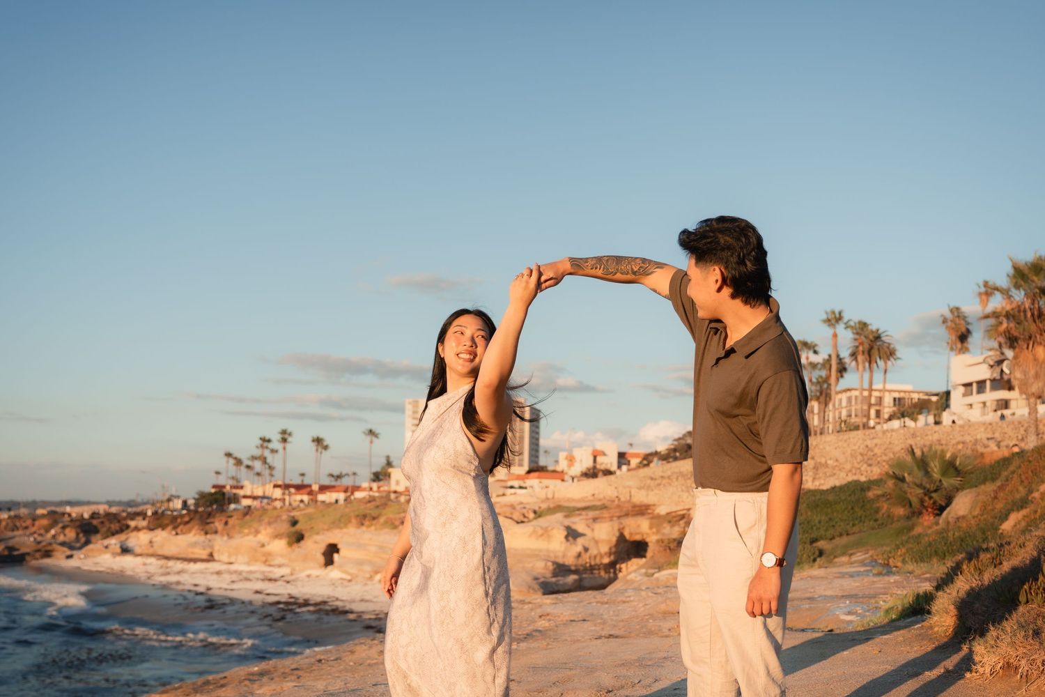 Couple dancing and twirling on a sandy coastal bluff at sunset with ocean waves, palm trees, and seaside buildings in the background.