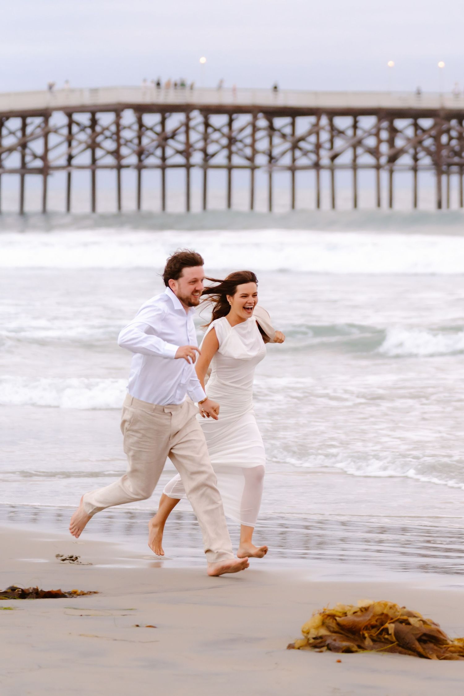 Laughing couple running barefoot on beach near a wooden pier, dressed in white and beige outfits by the ocean.