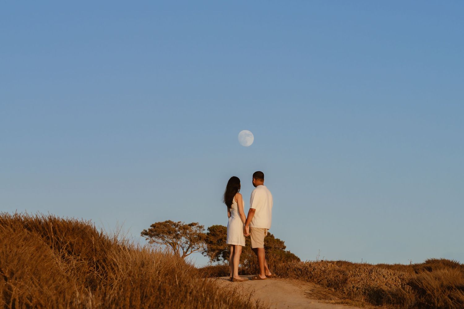 Couple holding hands on dirt path gazing at full moon rising over golden grass landscape.