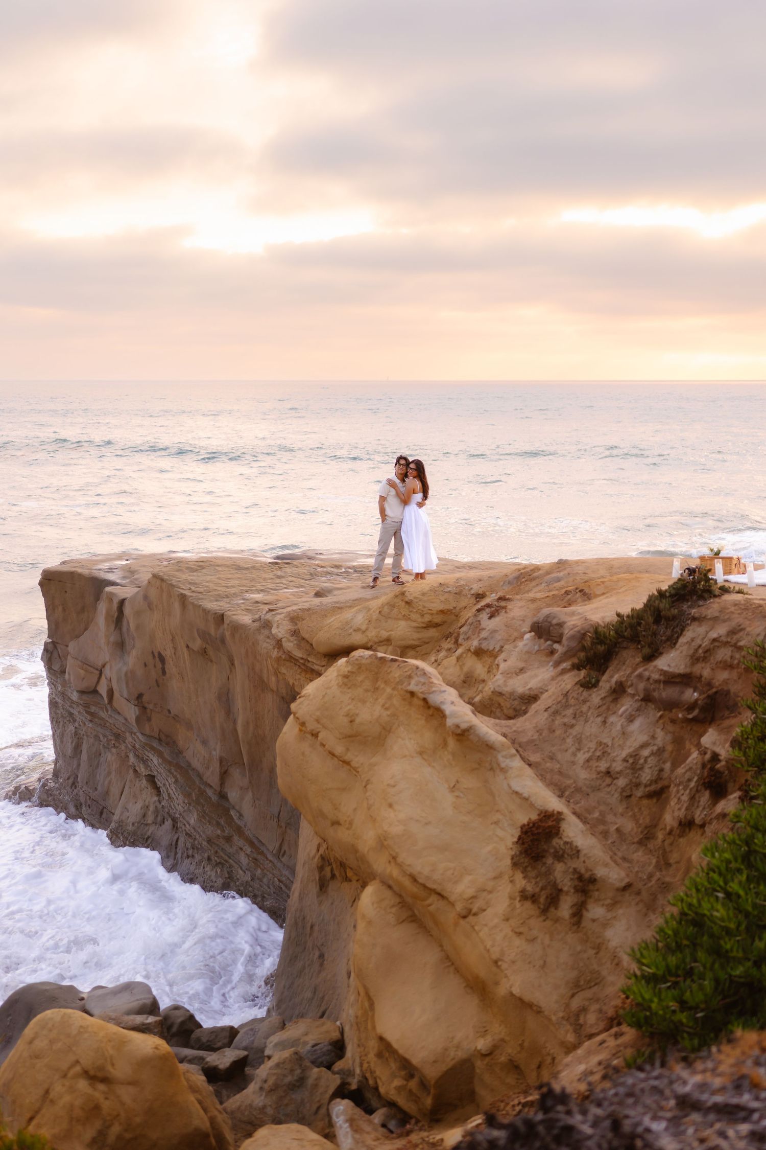 Couple embracing on dramatic rocky ocean cliff at sunset with crashing waves and golden sky. santa cruz cliffs san diego