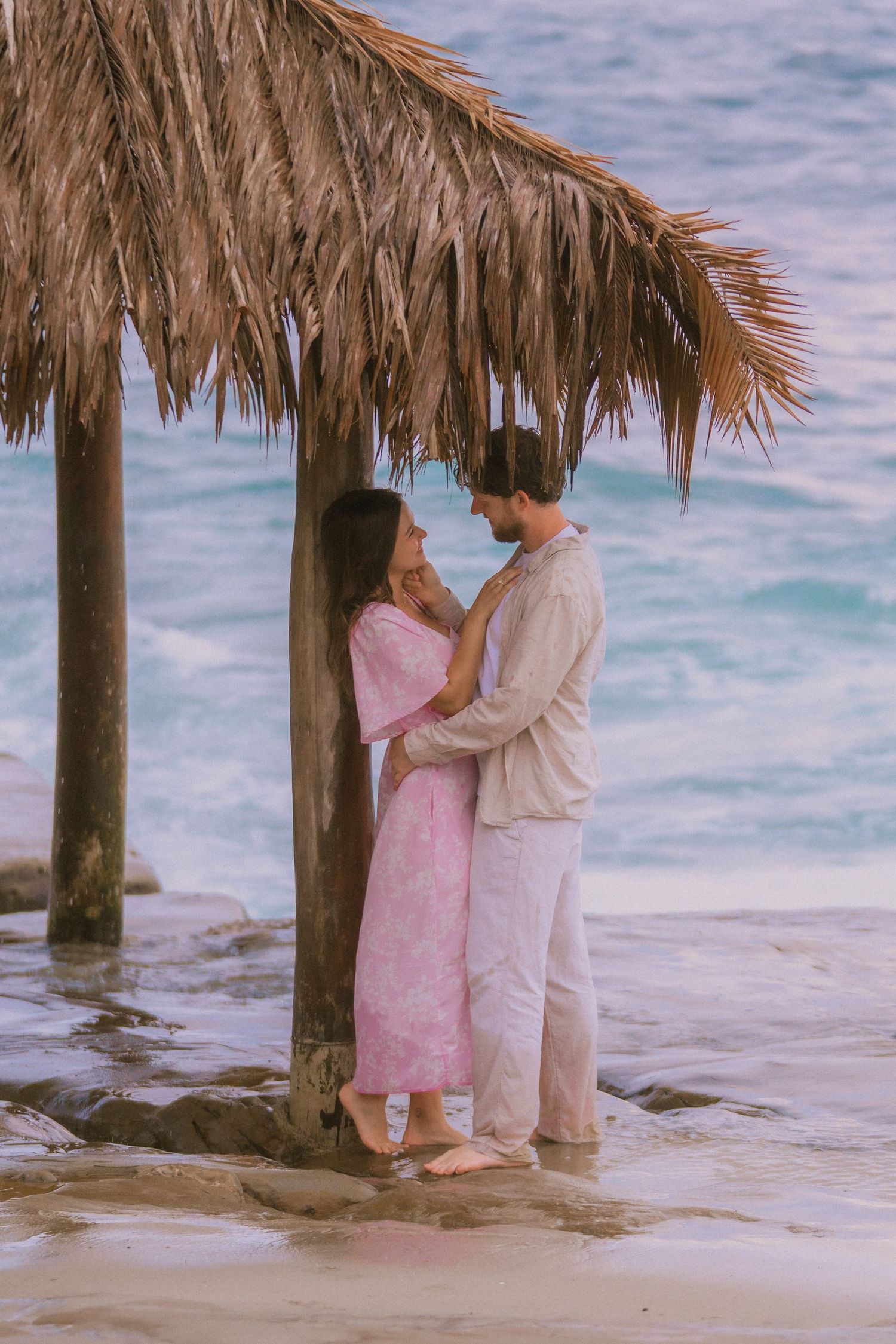 Romantic couple embracing under thatched palapa shelter on rocky ocean shore.