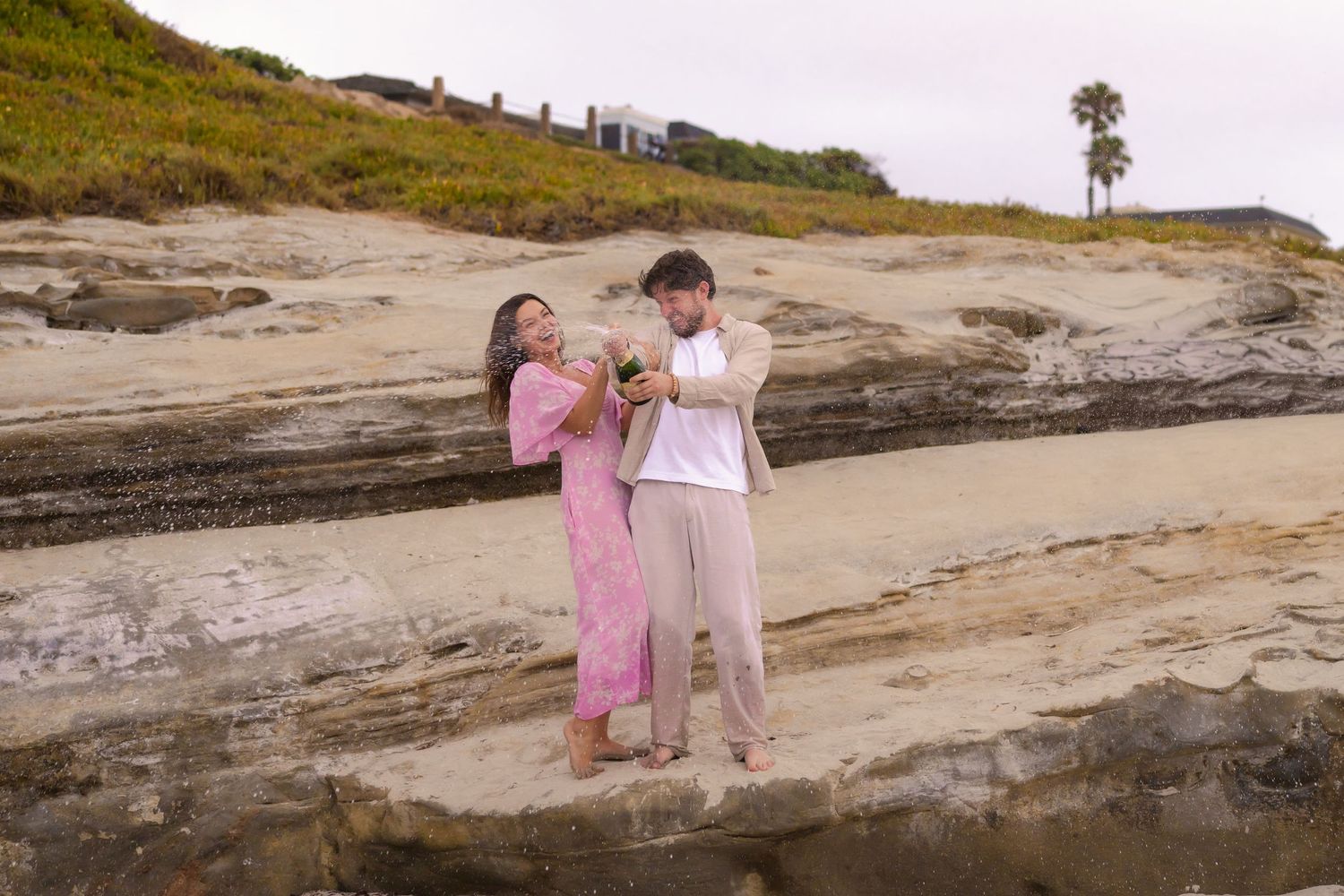 Happy couple examining engagement ring on rocky coastal cliffs at beach.