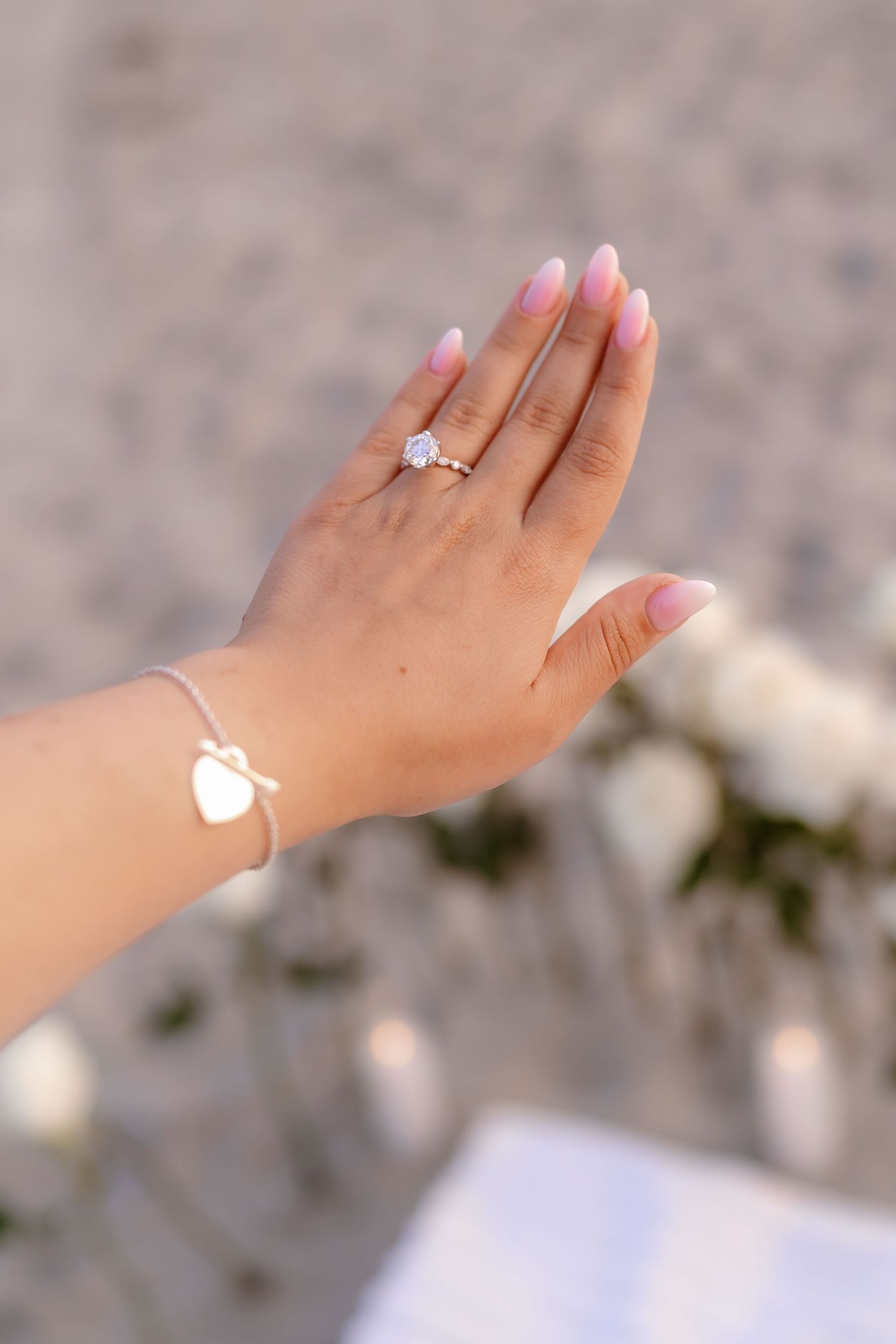 Close-up of engagement ring on woman's hand with pink ombre nails and heart bracelet.