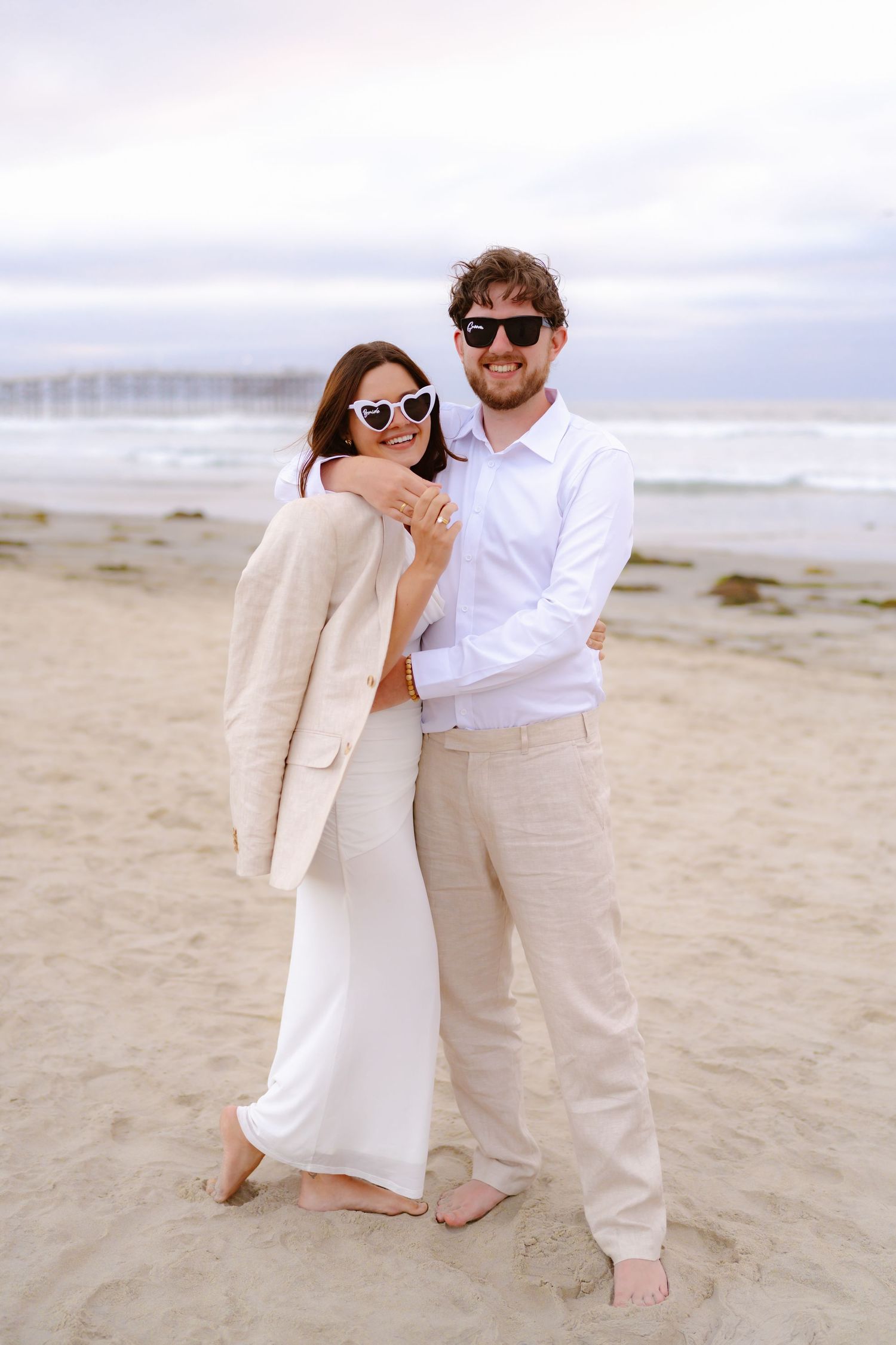 Happy couple posing on beach wearing heart-shaped sunglasses, woman in white dress with beige blazer, man in white shirt.