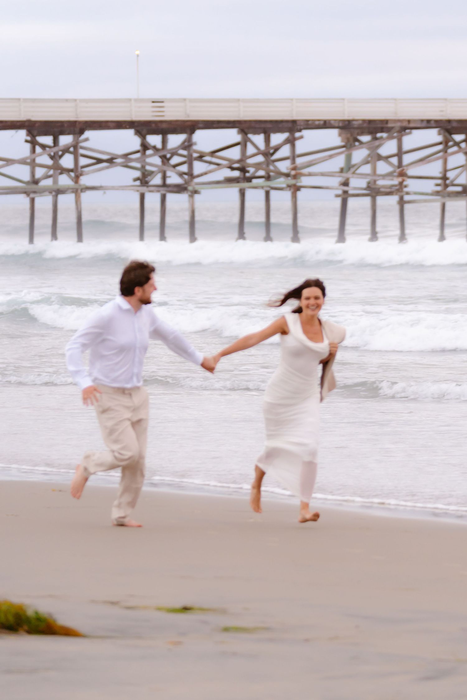 Joyful couple running hand in hand on a sandy beach near a pier, woman in white dress and man in white shirt.
