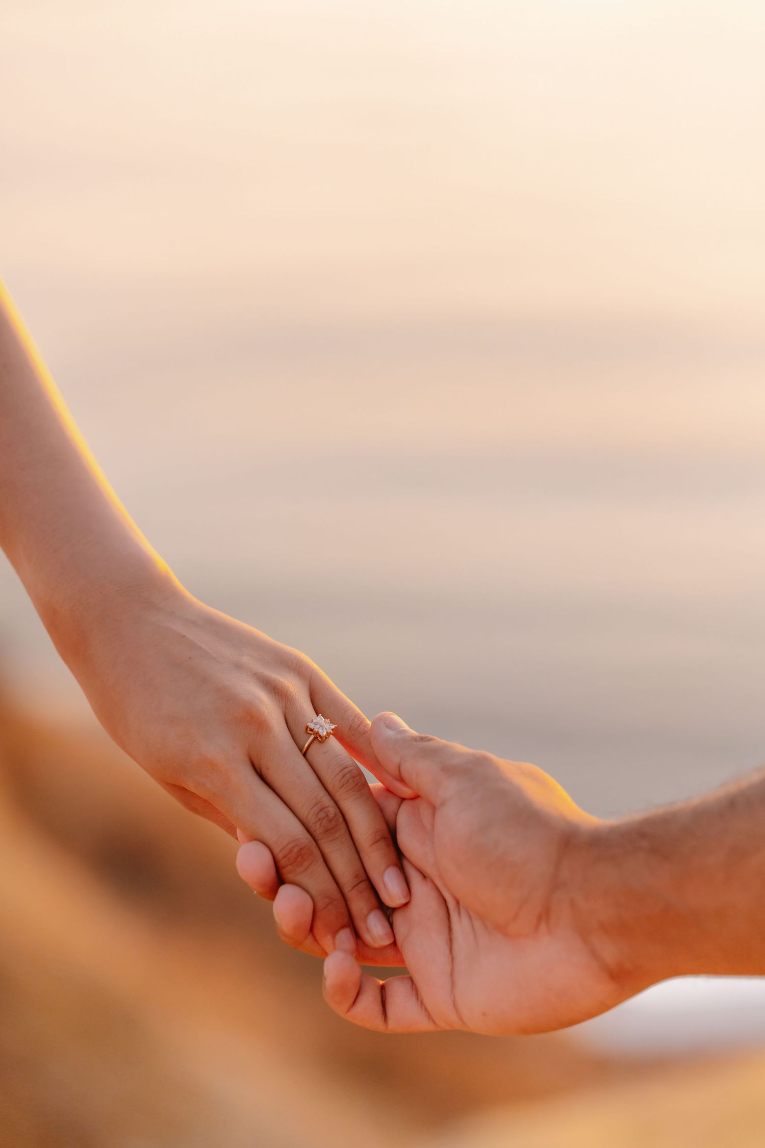 Close-up of couple holding hands showing engagement ring with soft pastel ocean background at sunset.
