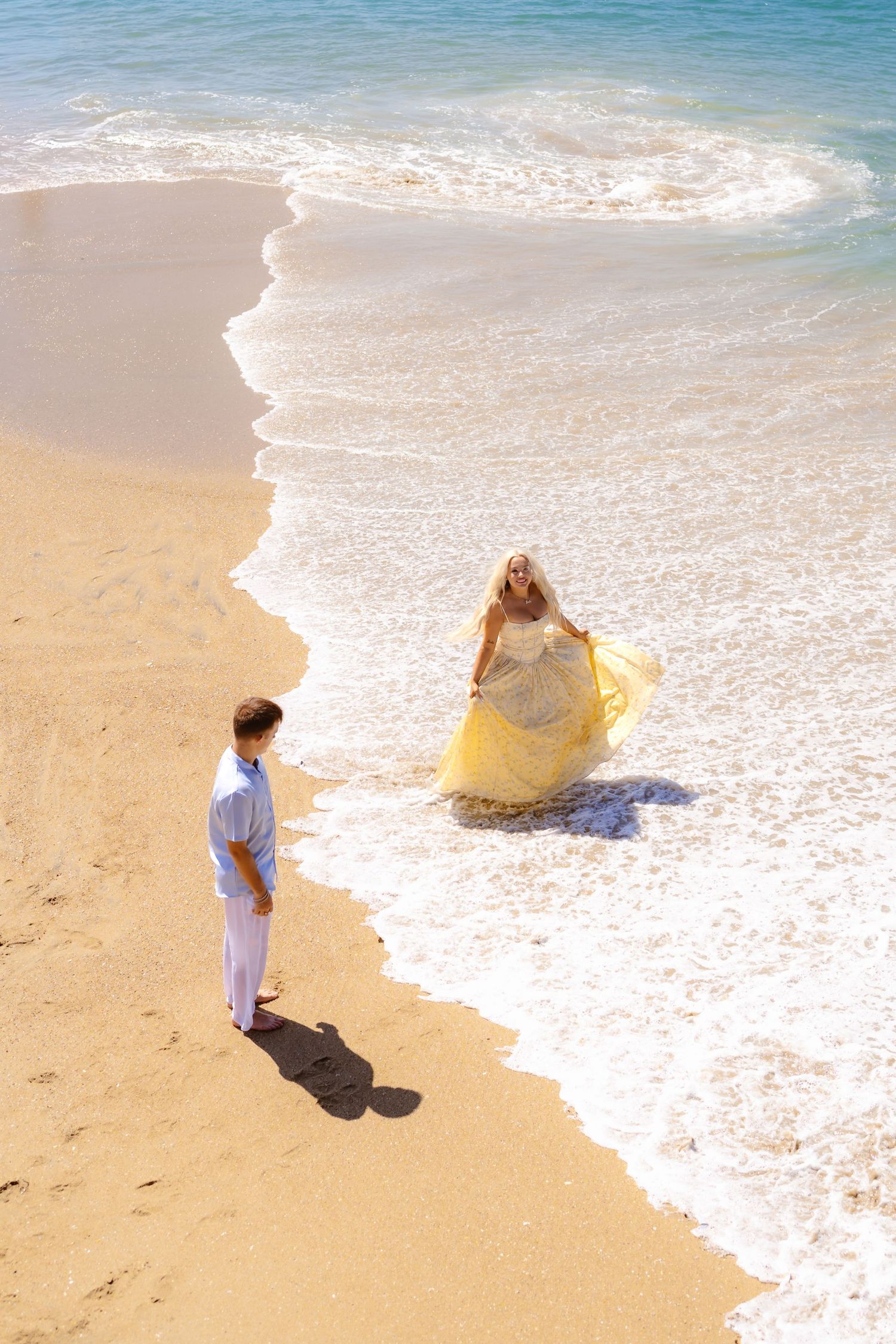 Woman in yellow gown walks toward man in white on sunny beach with ocean waves.