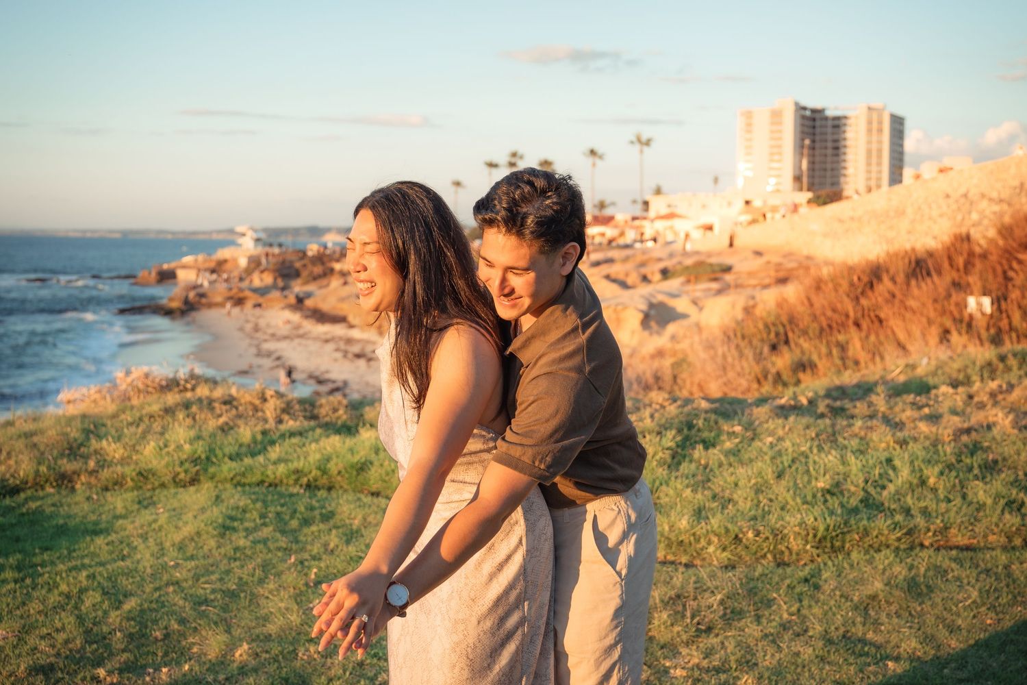 Smiling couple embracing on a grassy coastal hillside at sunset with ocean waves, a sandy beach, and city buildings in the background.