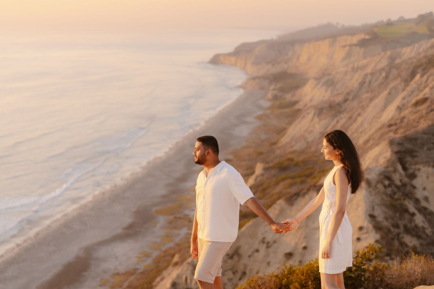 Couple in white outfits holding hands on coastal cliffs overlooking scenic beach at golden sunset. Torrey Pines