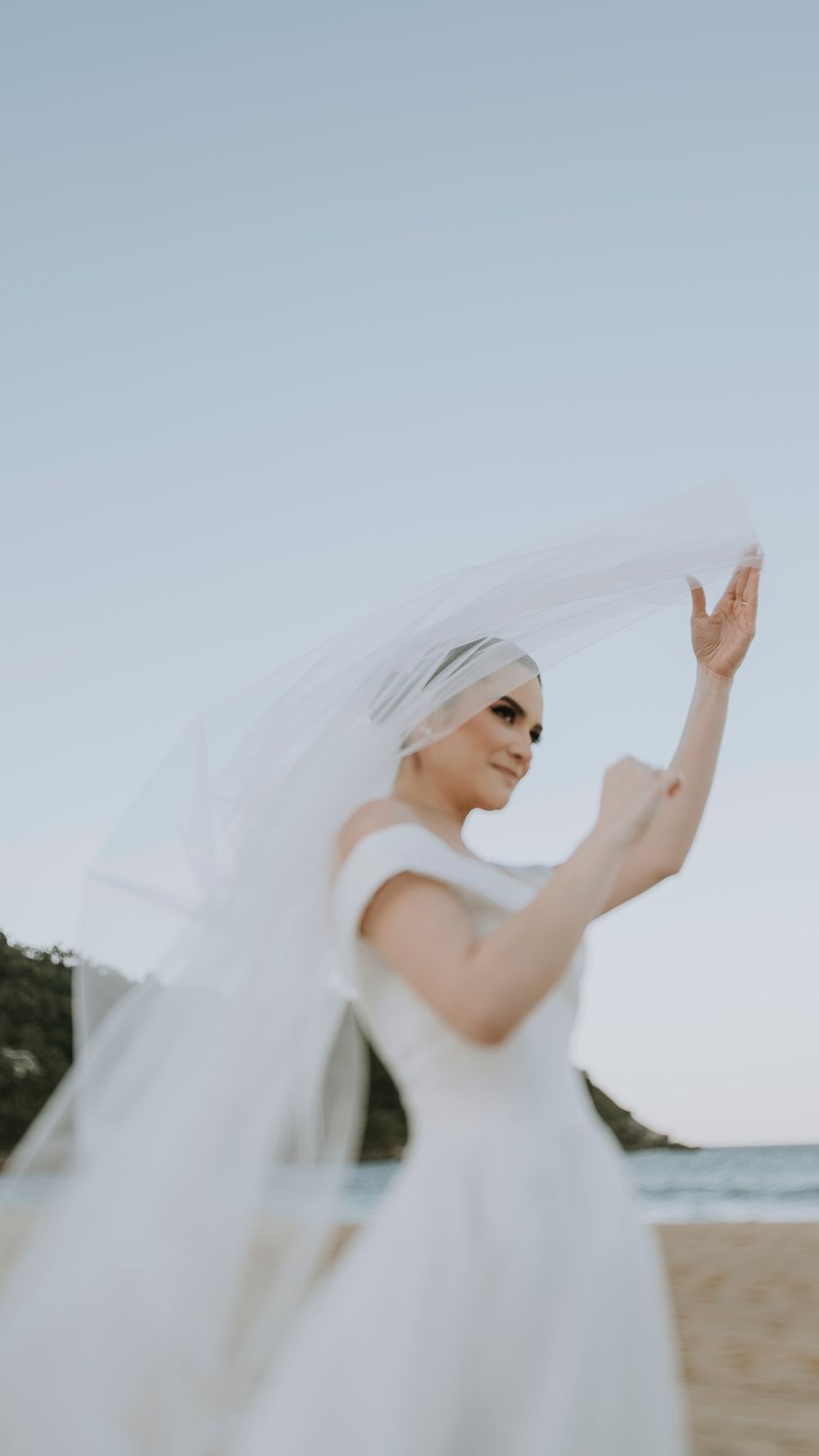 Bride in a white gown and veil poses gracefully on a beach against a clear sky.