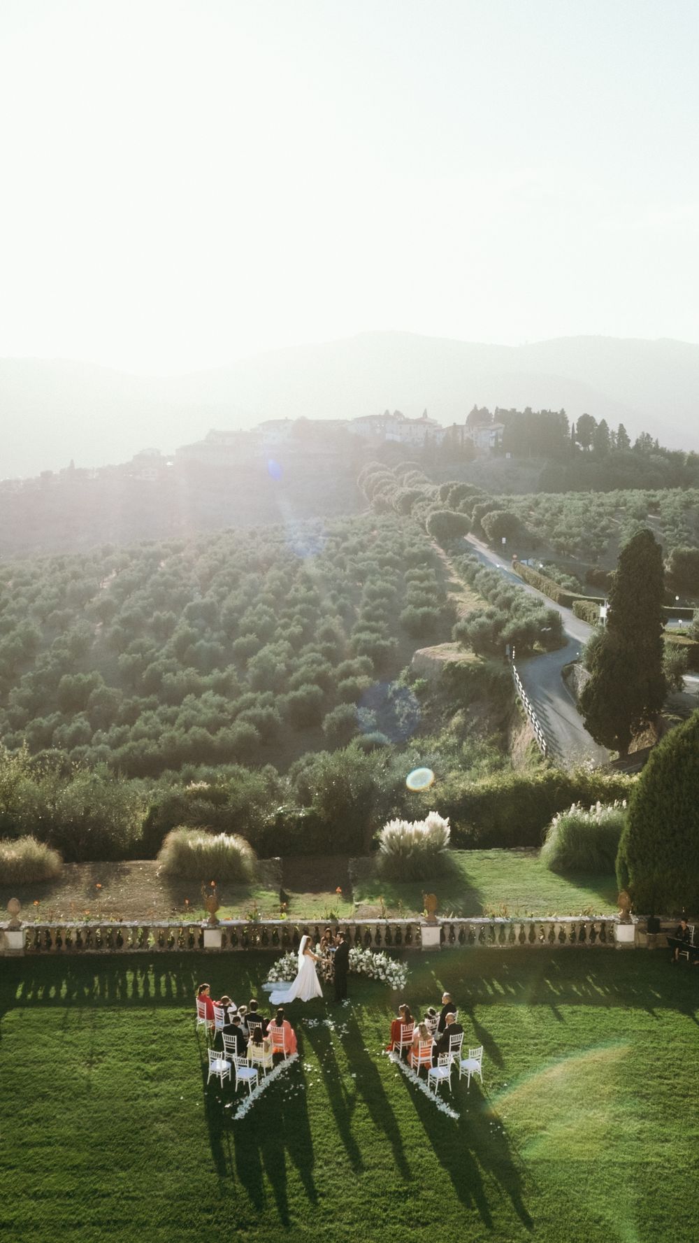 Aerial view of intimate outdoor wedding ceremony on lush green lawn surrounded by Tuscan landscape.