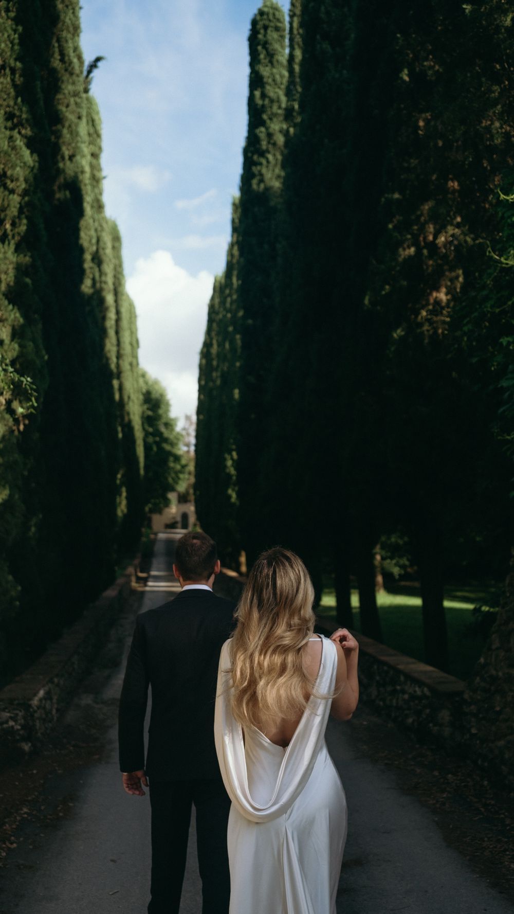 Wedding couple walking hand-in-hand down a tree-lined path, bride in white gown and groom in black suit.