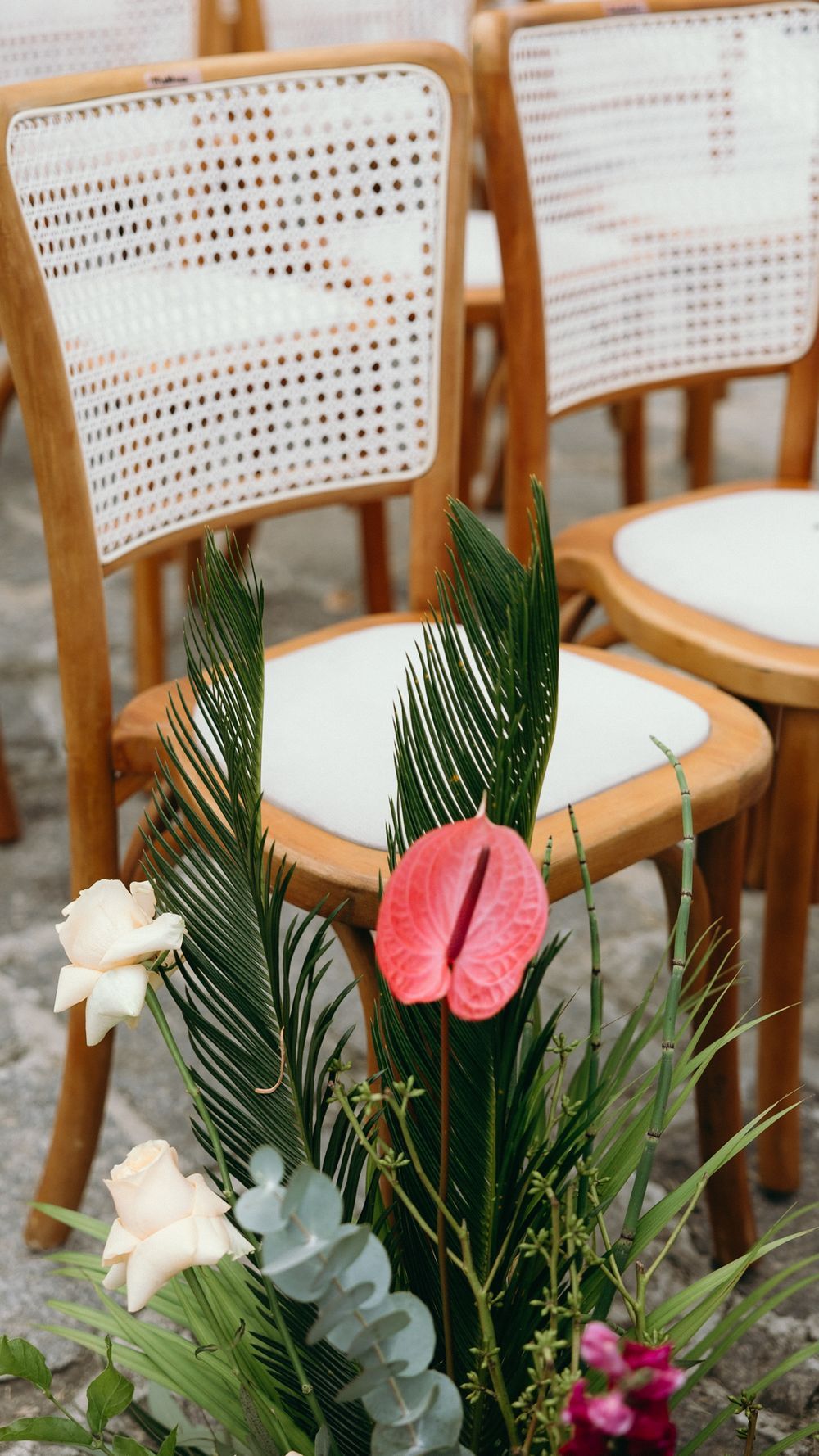 Wooden cane-back ceremony chairs adorned with tropical floral arrangement featuring pink anthurium and palm leaves.