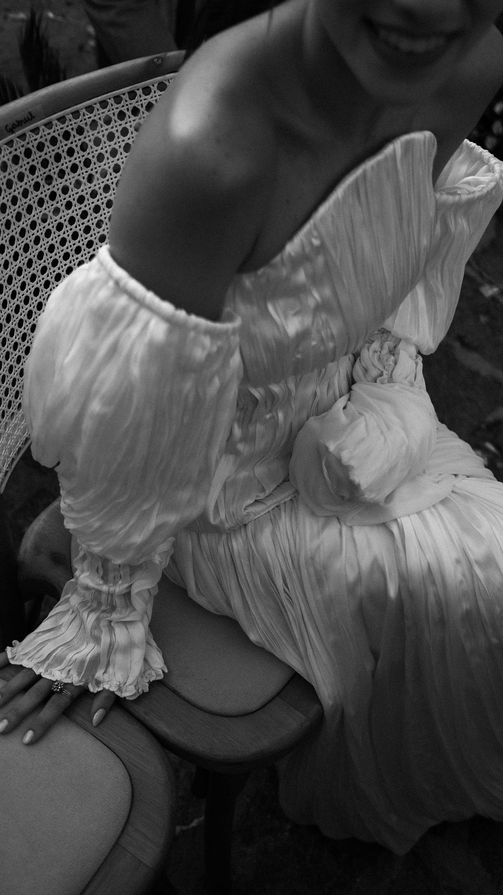 Black and white photo of smiling bride in elegant off-shoulder pleated white wedding dress seated in chair.