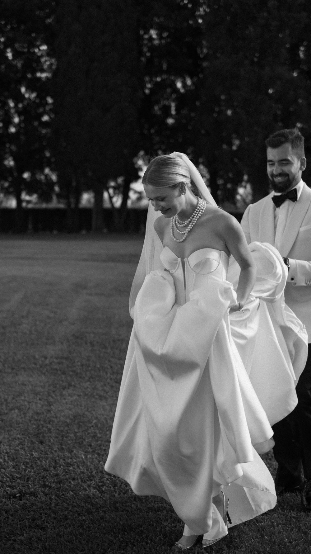 Black and white photo of laughing bride lifting gown hem as groom smiles behind her outdoors.