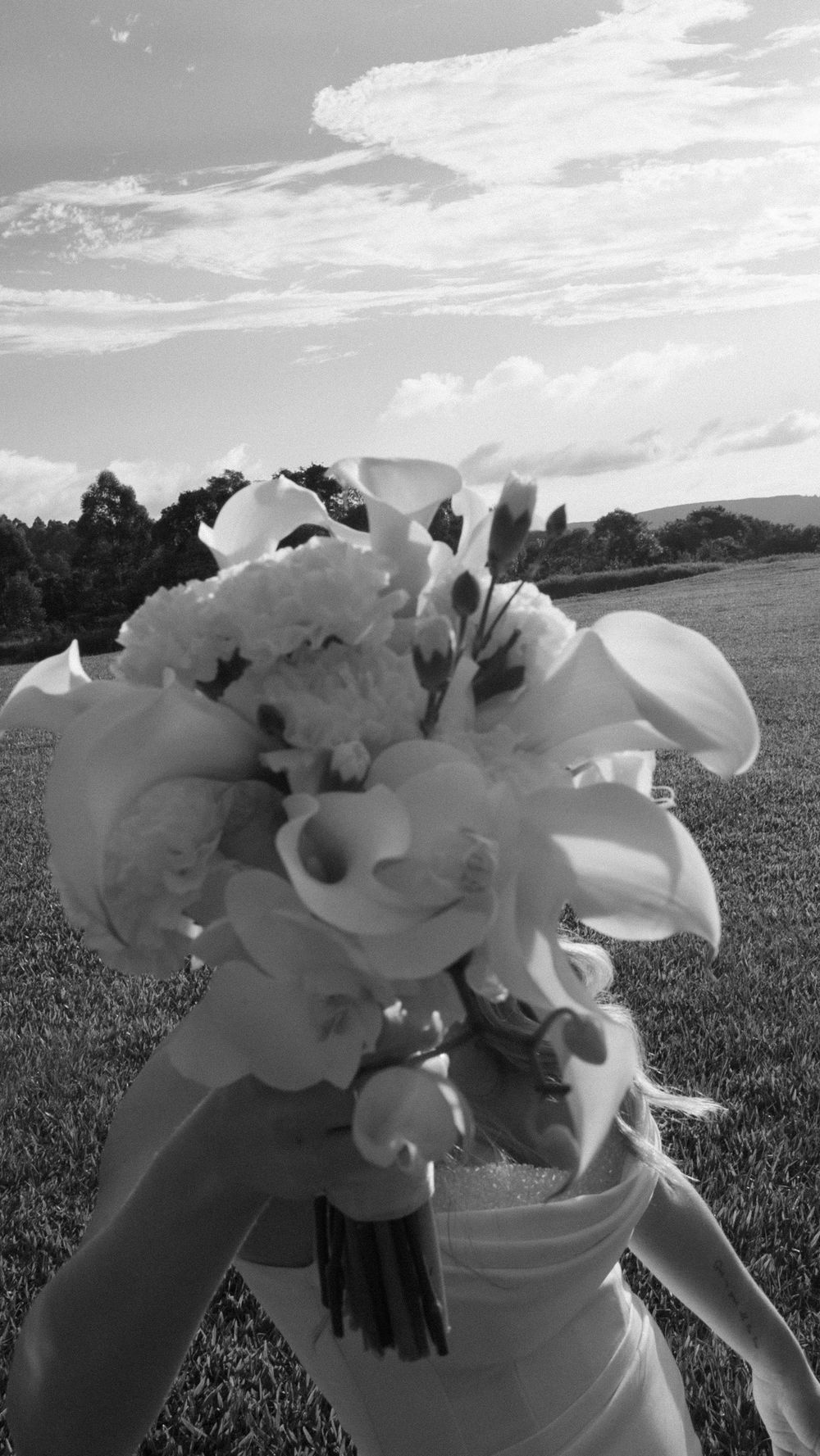 Bride holds a bouquet of calla lilies and roses against a cloudy sky in black and white.