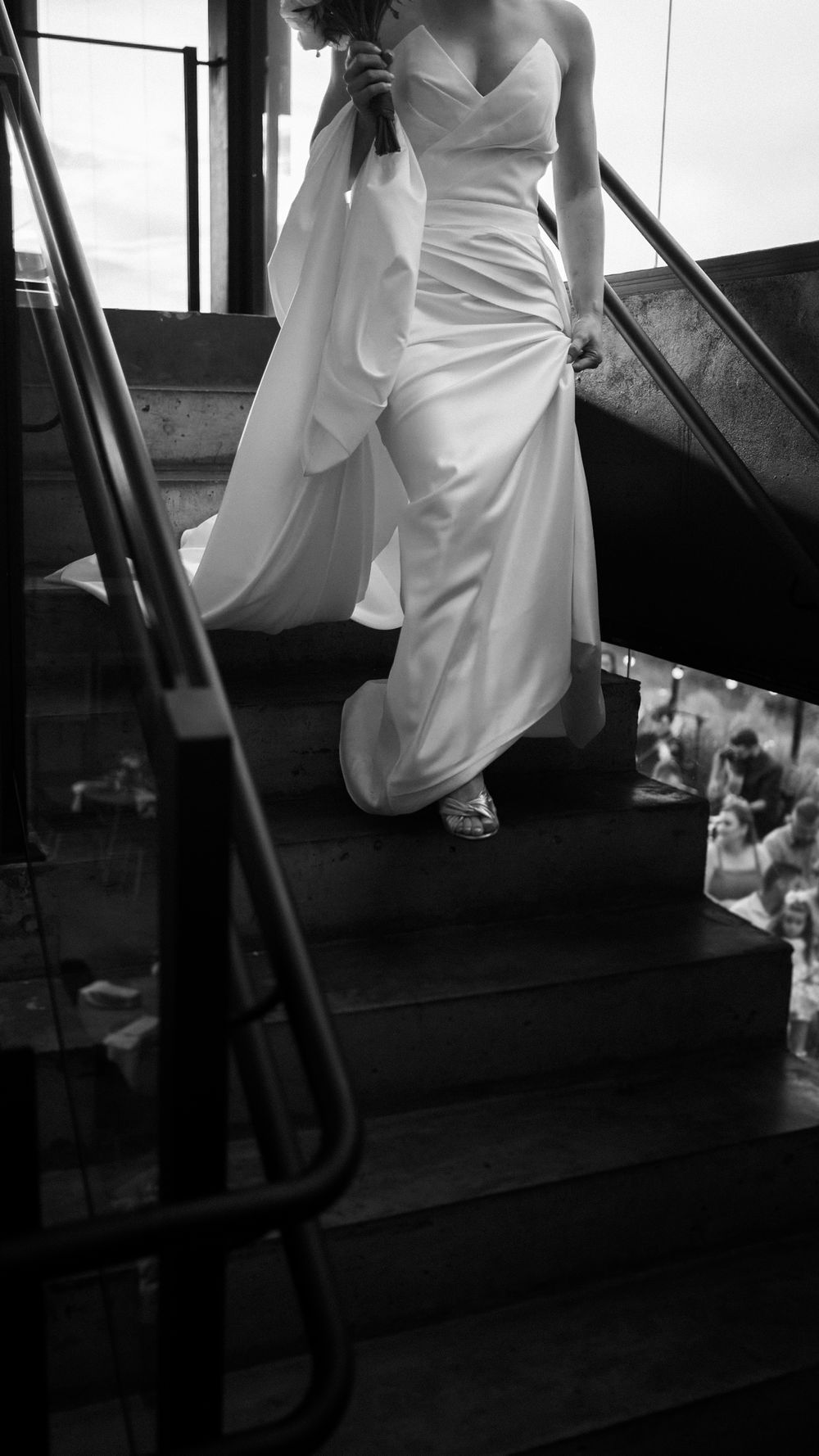 Black and white photo of a bride in a sleek white gown descending grand staircase holding a bouquet.