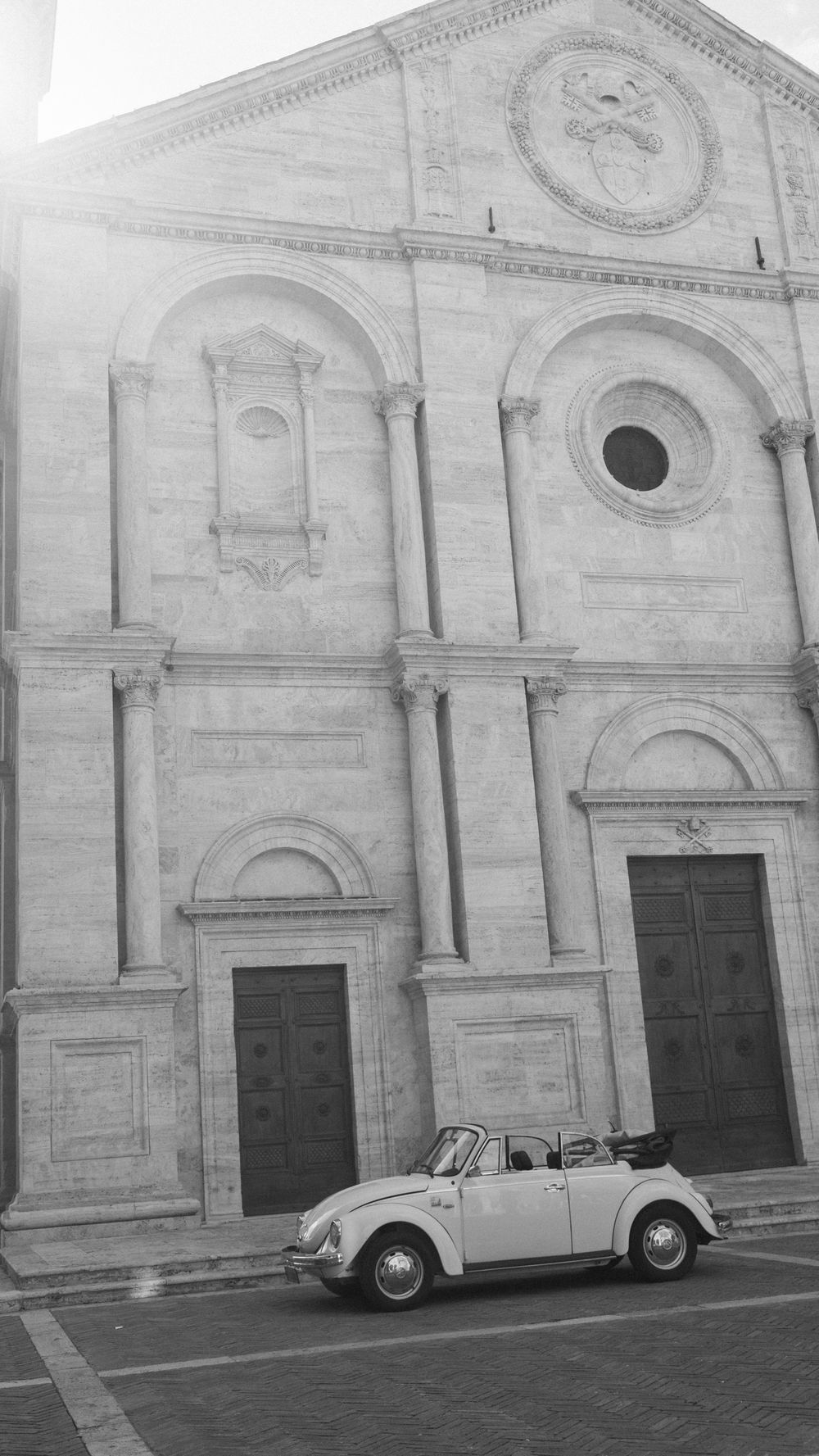Black and white photo of a vintage VW Beetle convertible parked in front of a grand Renaissance church facade.