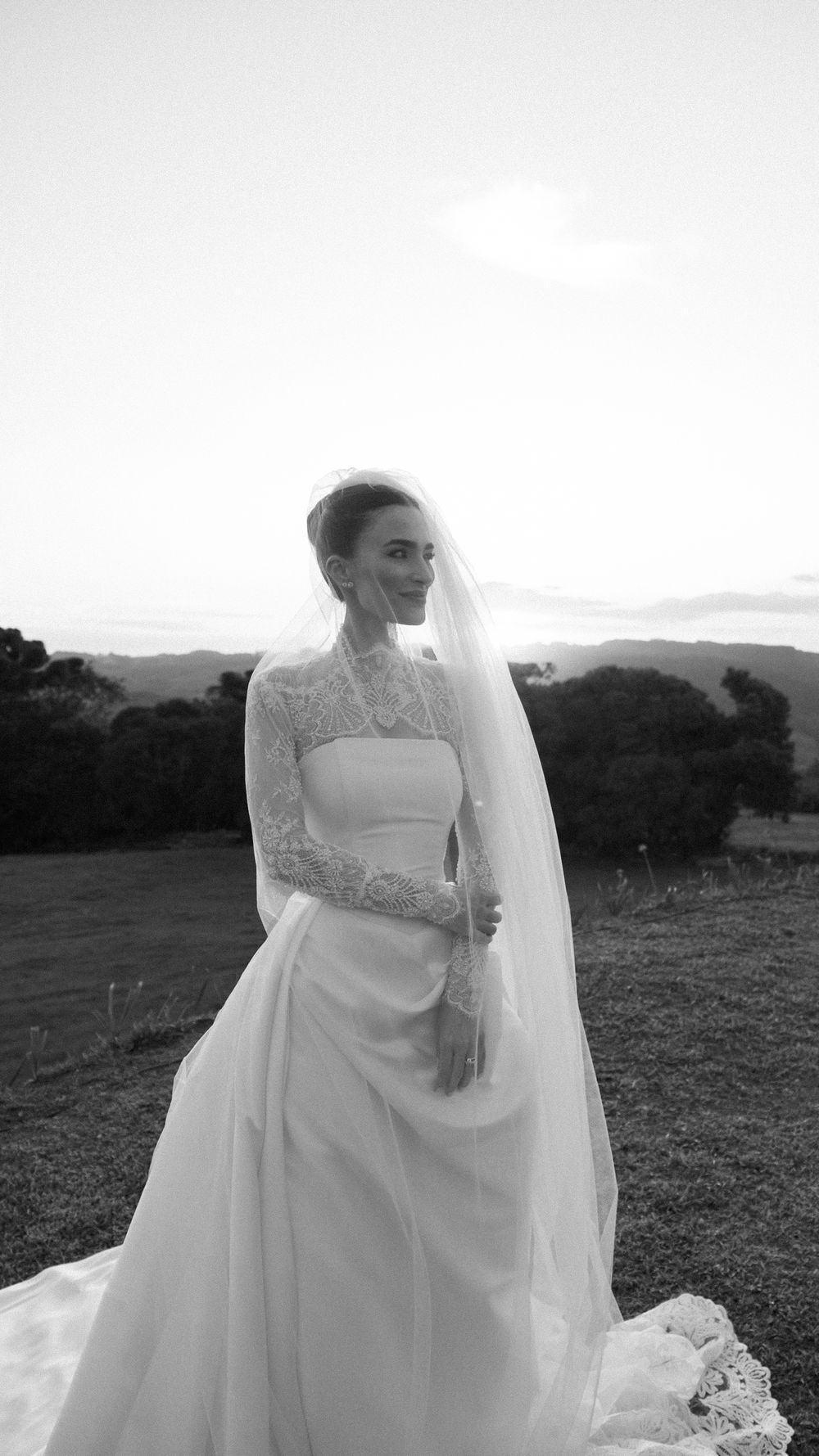 Elegant bride in lace long-sleeve gown and veil at sunset in black and white.