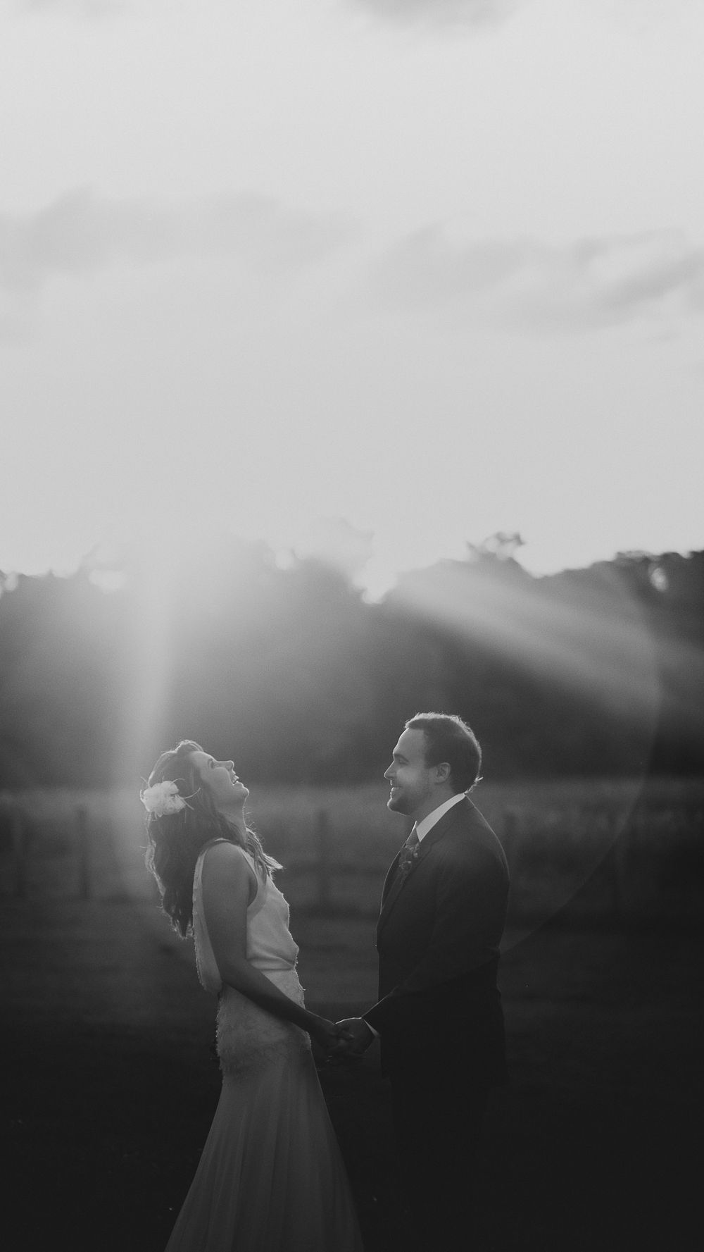 Black and white romantic photo of bride and groom holding hands with sunrays shining behind them.