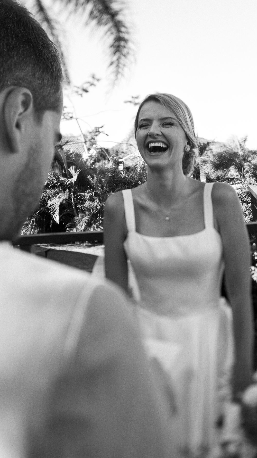 Laughing bride in white dress shares a joyful moment with groom outdoors in black and white.