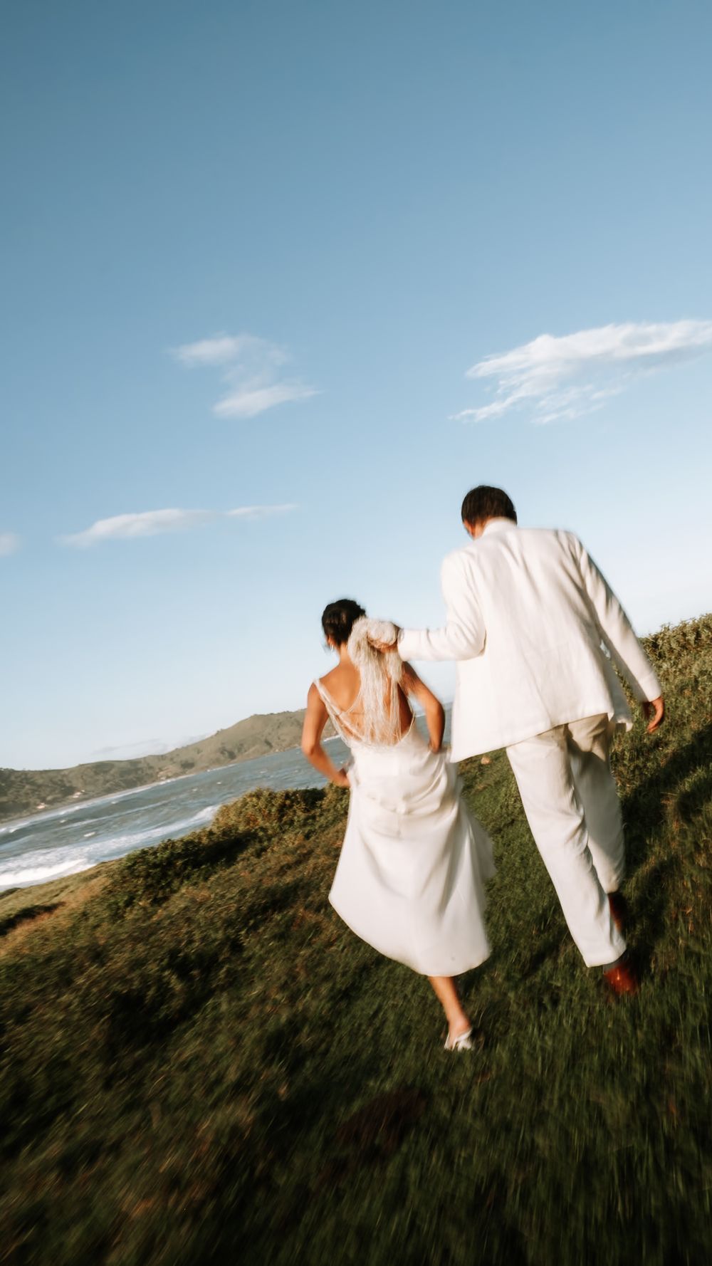 Bride and groom walking hand in hand through lush green coastal hillside with ocean and mountains in background.