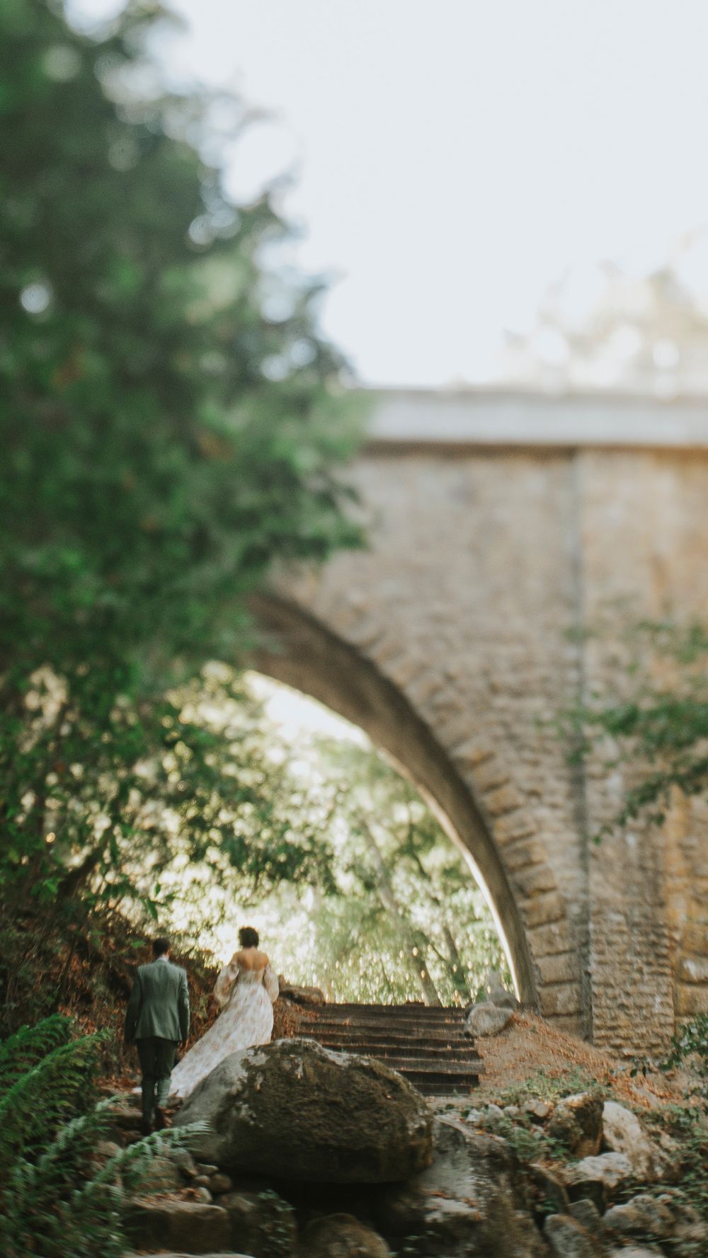 Bride and groom stand beneath a stone arch bridge surrounded by lush green trees at golden hour.