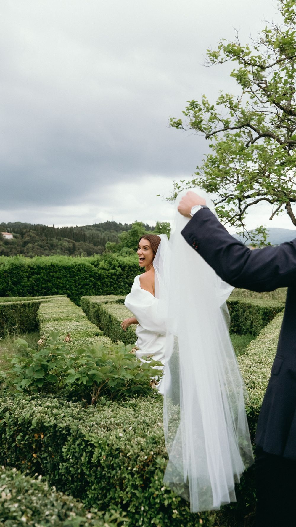 Groom lifting bride's veil as she laughs joyfully in a lush green garden on their wedding day.