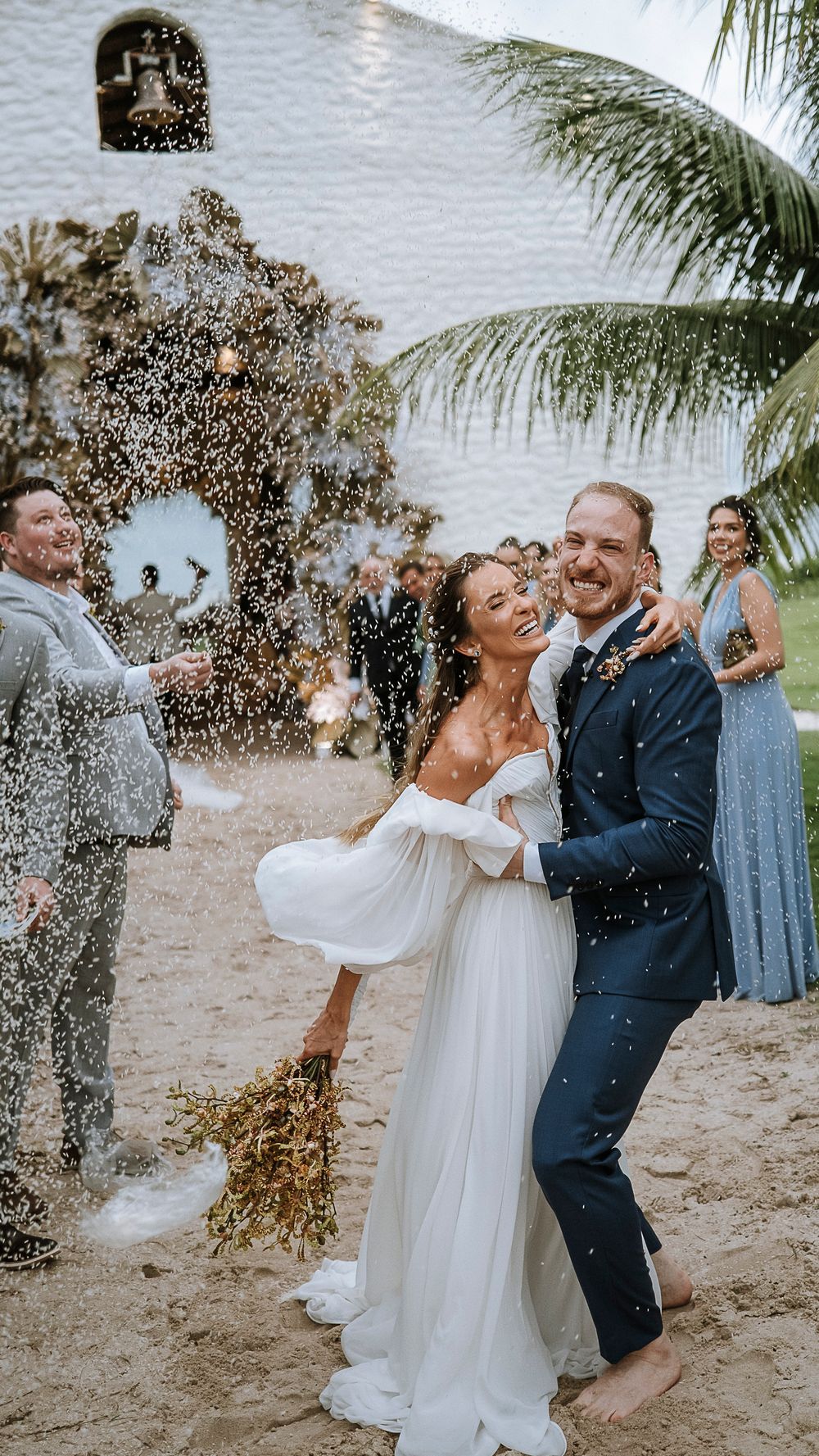 Joyful couple celebrating their beach wedding exit with confetti shower surrounded by guests near a white chapel.