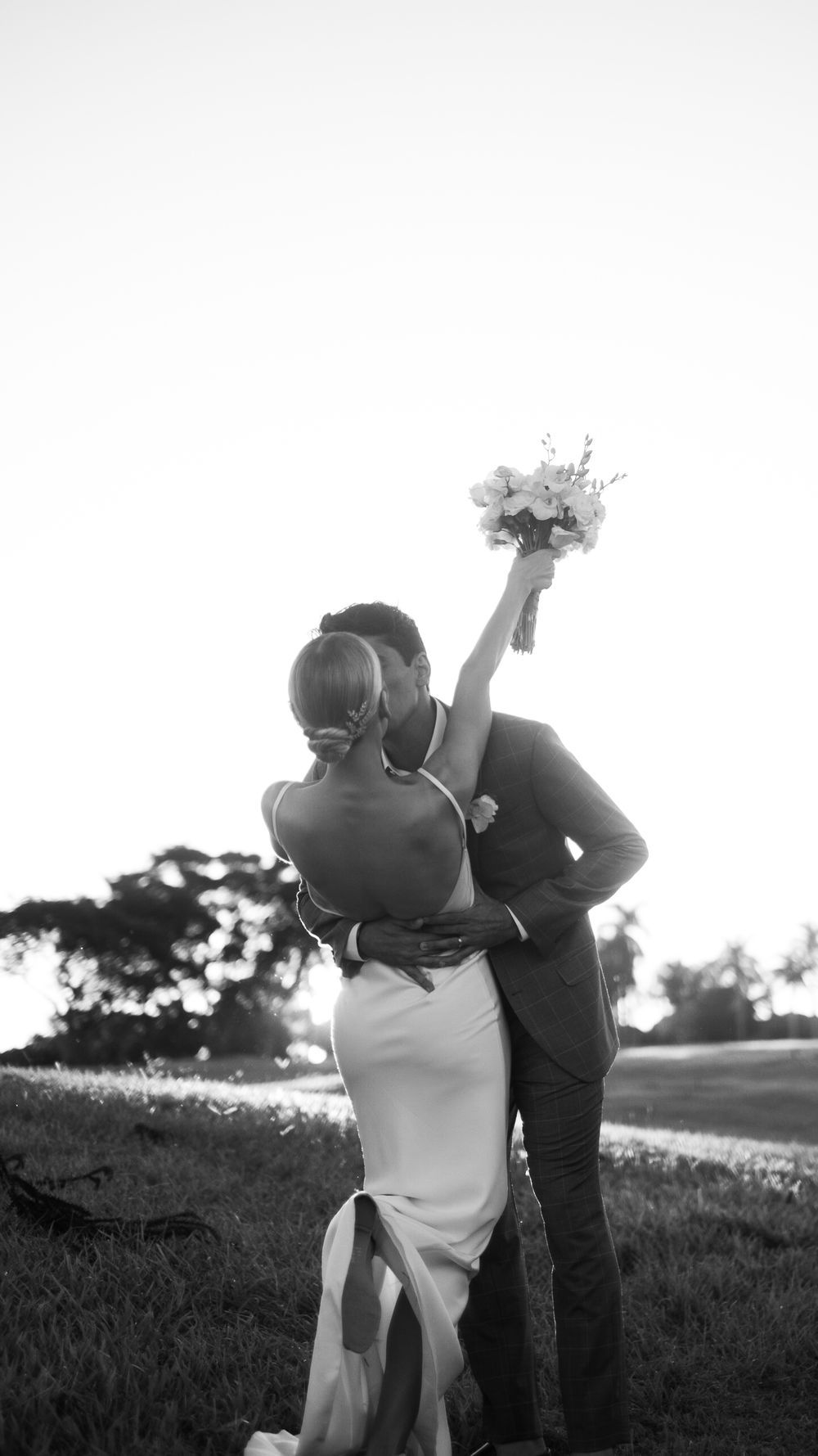 Black and white photo of groom kissing bride holding floral bouquet at sunset outdoor wedding.