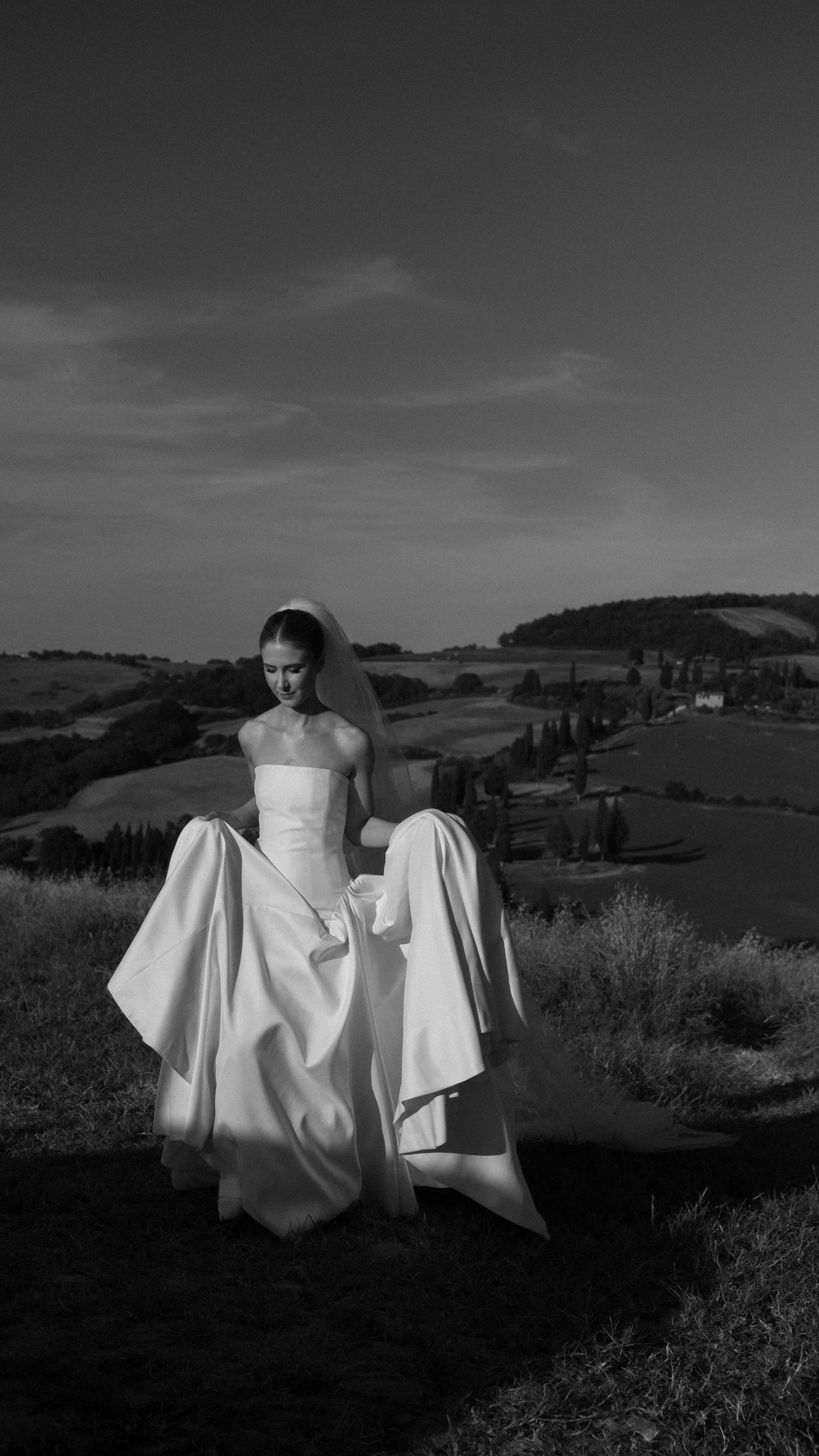 Black and white photo of a bride in a strapless gown and veil, holding her dress on a scenic hilltop.