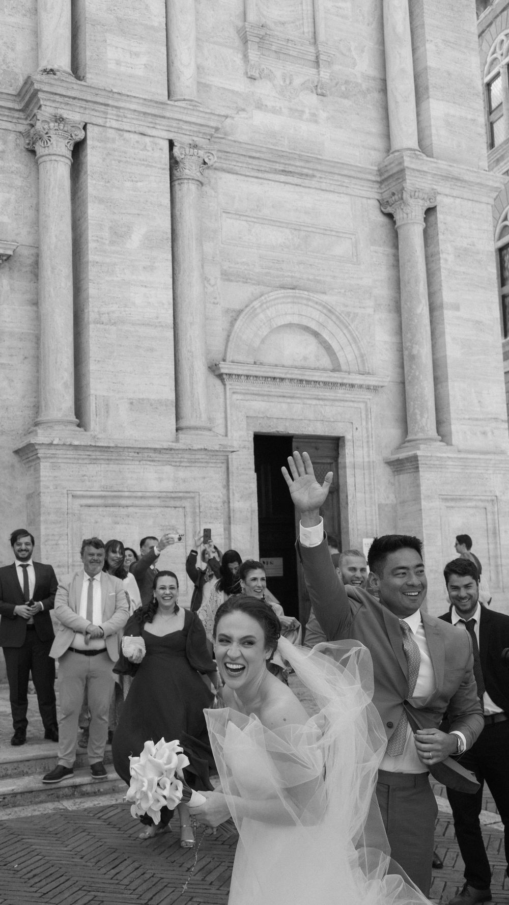 Joyful newlyweds celebrate with guests outside a grand Italian church, bride holding bouquet and groom waving.