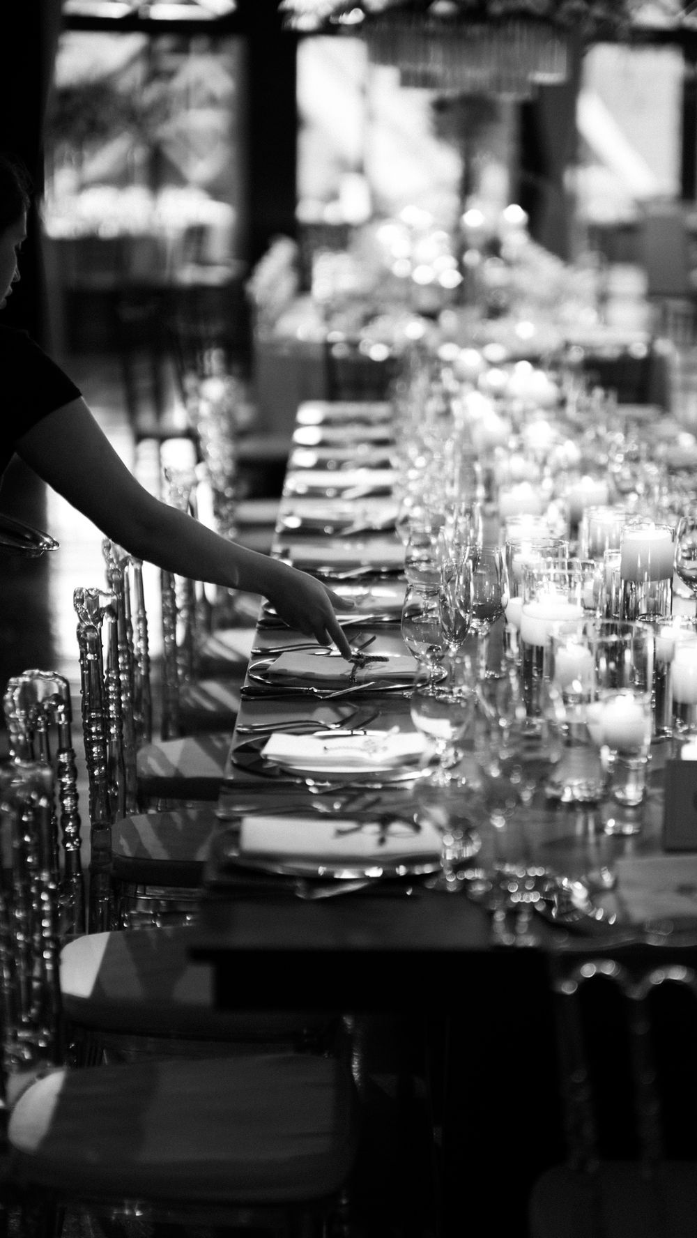 Black and white image of a server setting a long elegantly arranged wedding reception dining table.