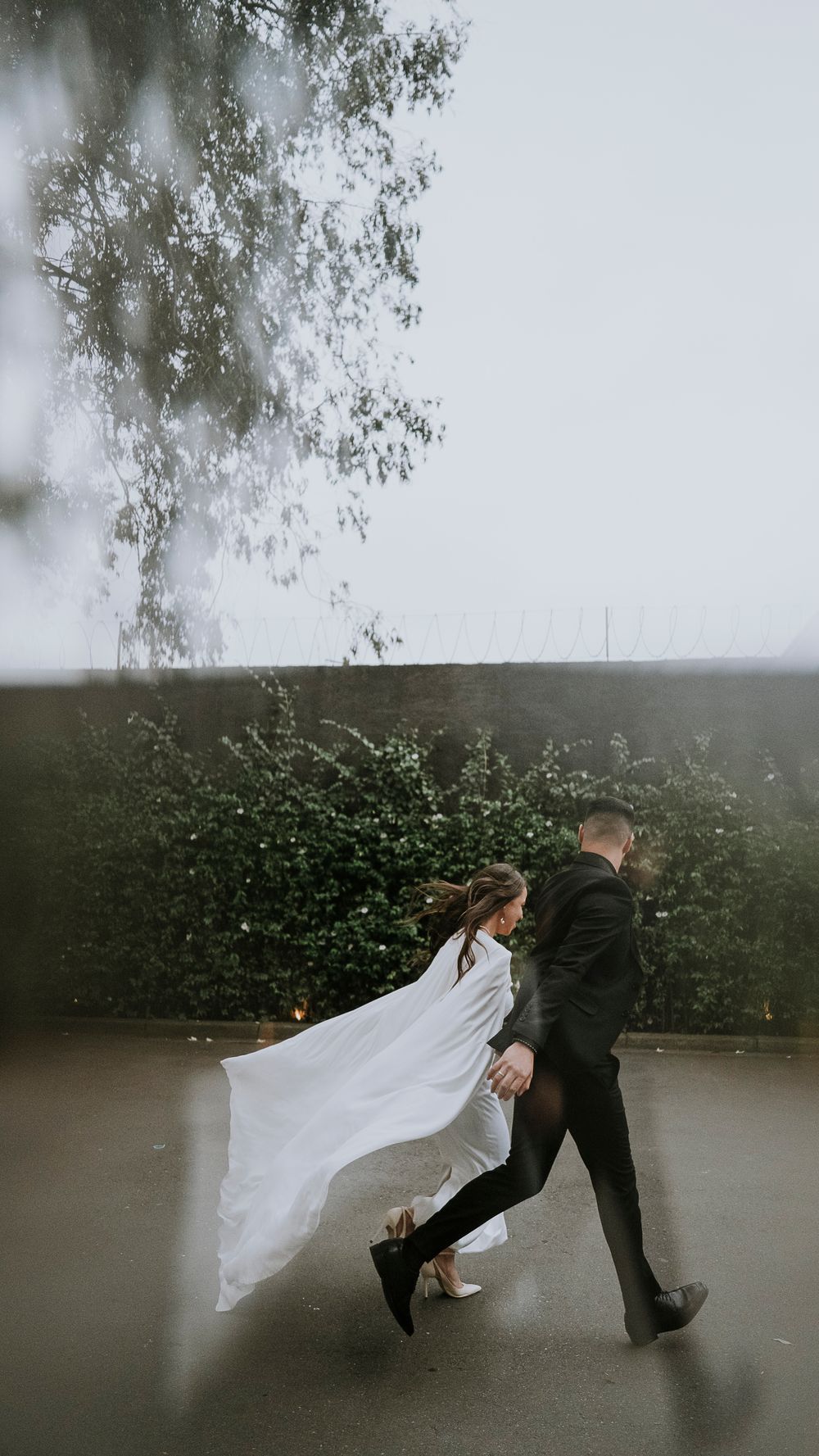 Bride and groom walking hand in hand with wedding veil flowing in the wind against a lush green hedge.