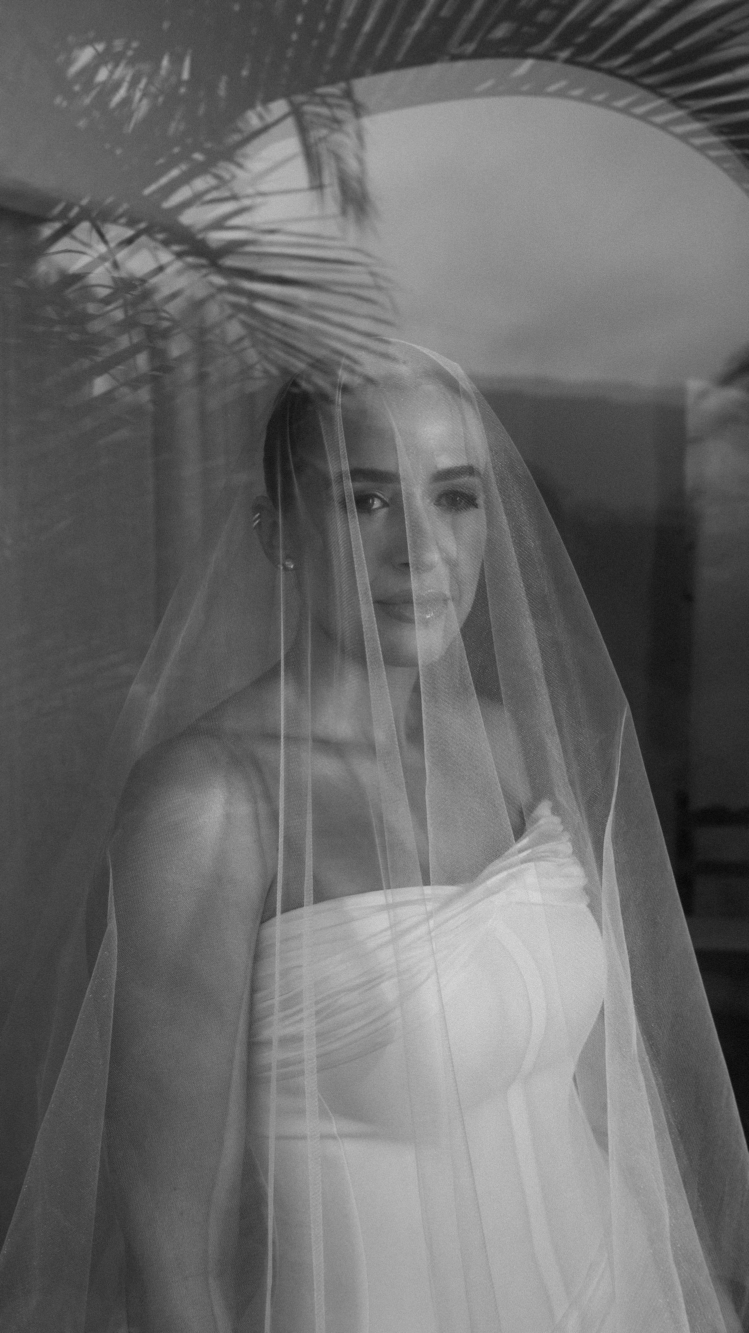 Elegant bride in strapless gown and long veil poses beneath palm fronds in black and white portrait.