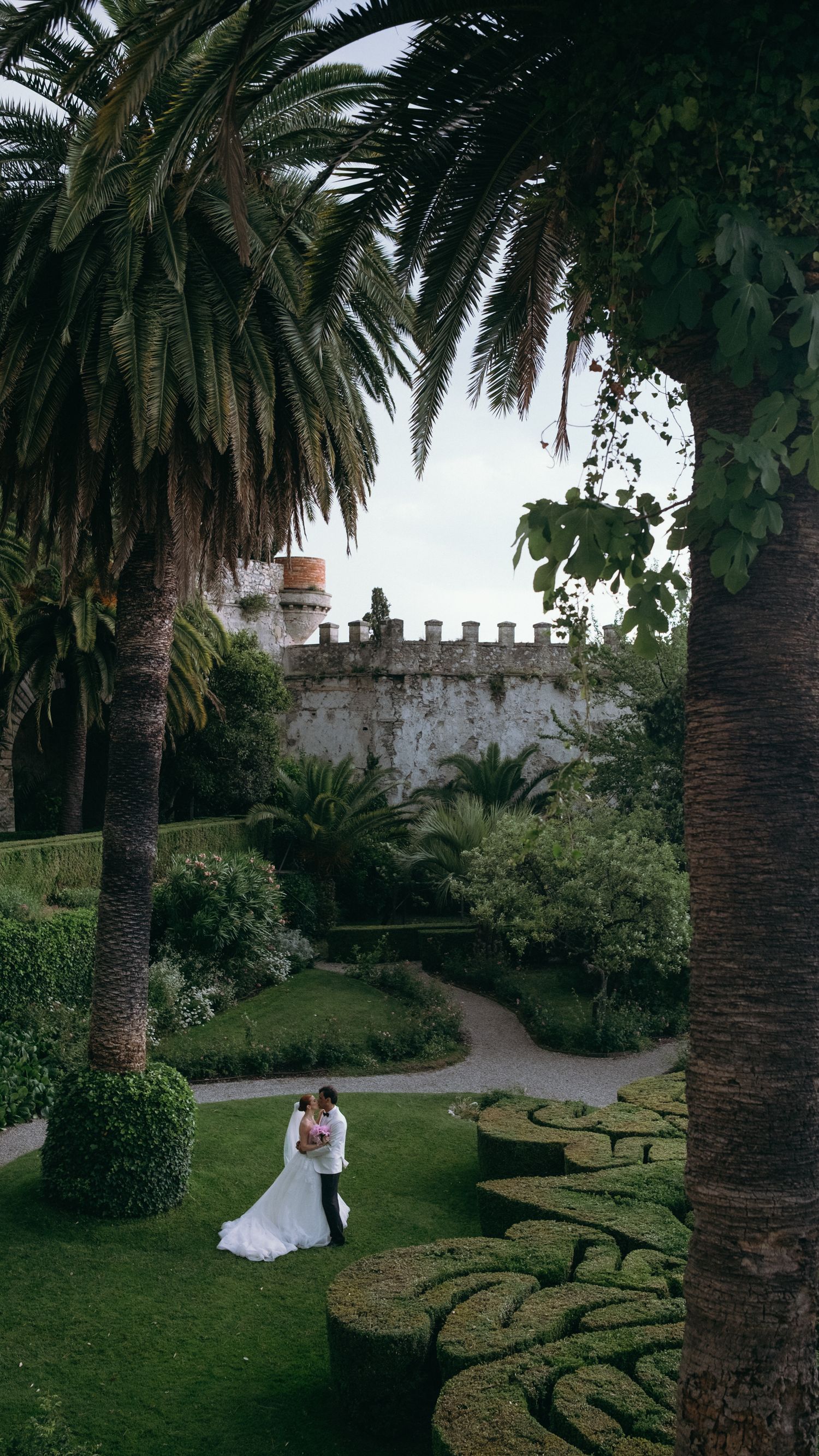 Bride and groom share a romantic moment in a lush garden with palm trees and historic castle walls.