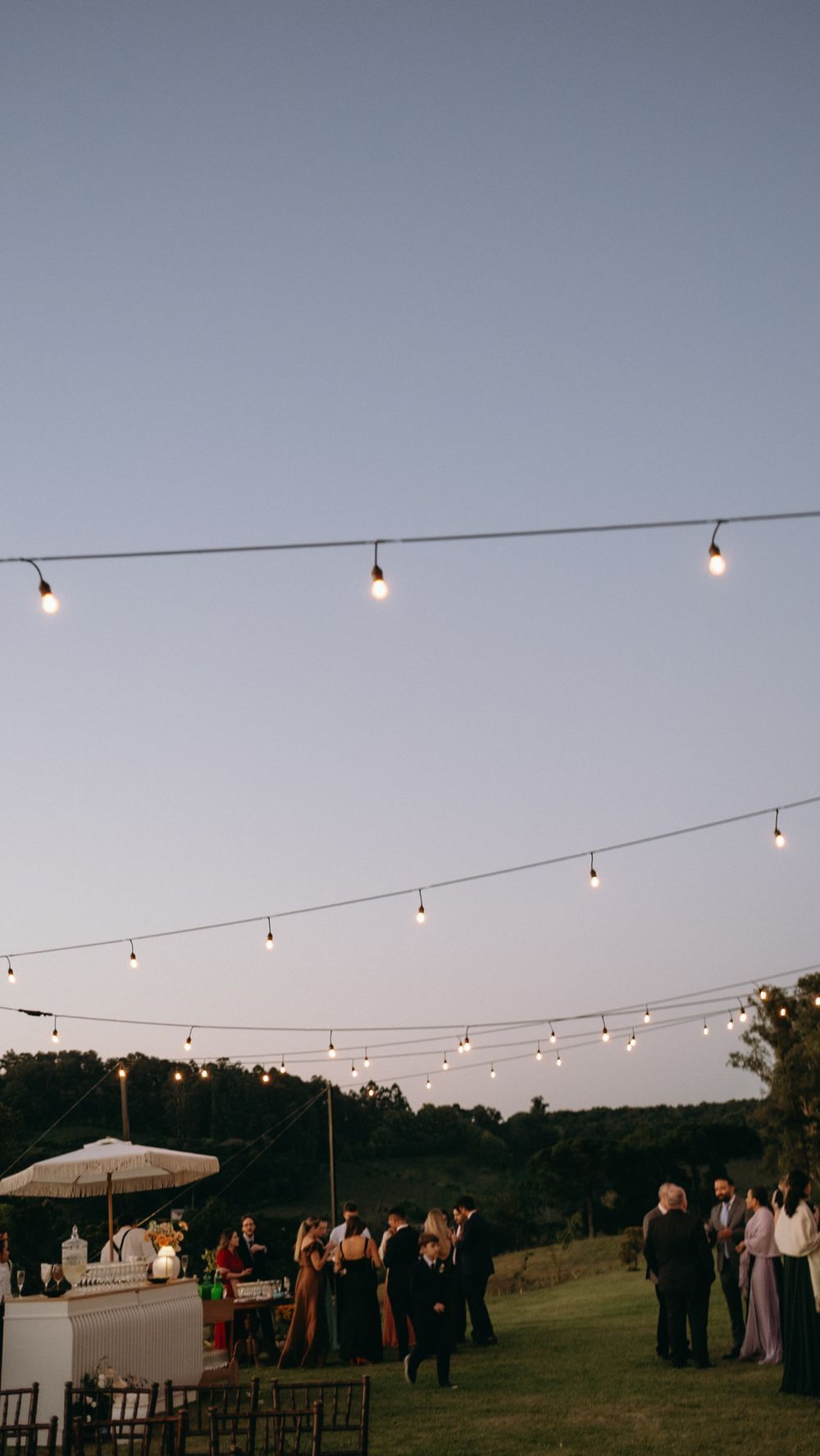 Outdoor wedding reception at dusk with string lights and guests on green lawn.