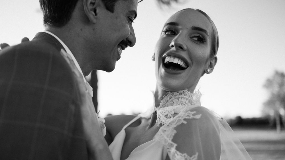 Black and white photo of laughing bride in lace wedding dress with groom at outdoor ceremony.