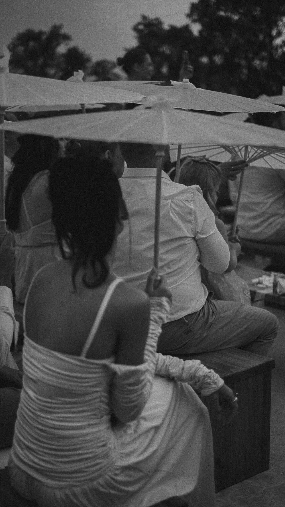 Black and white photo of wedding guests seated under parasol umbrellas at outdoor event.