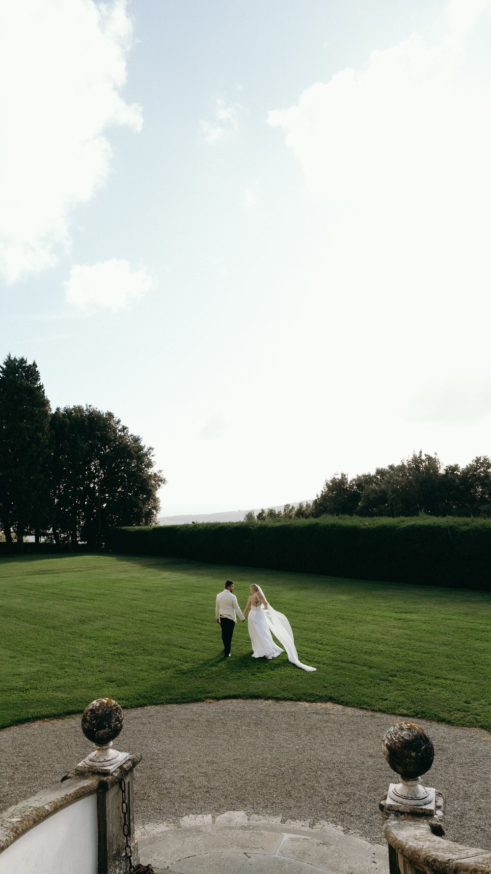 Bride and groom walk hand in hand across lush green lawn of elegant Italian estate.