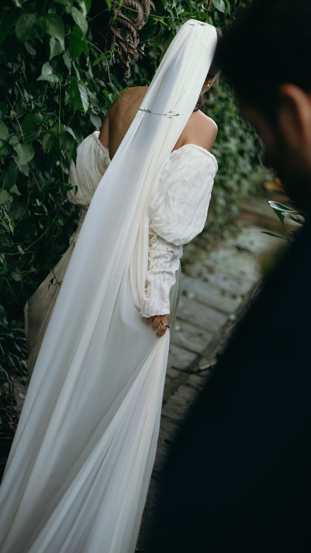 Bride in white off-shoulder gown with long veil walks through lush green garden pathway on wedding day.