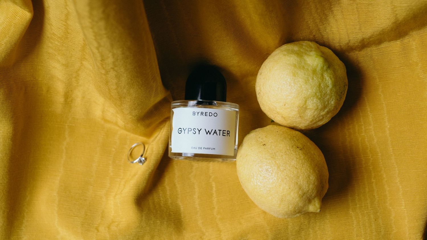 Byredo Gypsy Water perfume bottle beside two fresh lemons on a golden yellow linen cloth.