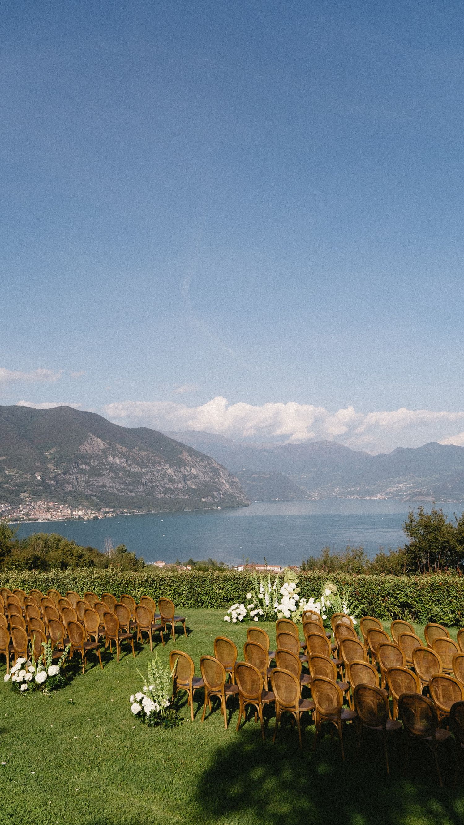 Elegant outdoor wedding ceremony chairs arranged on a green lawn overlooking a scenic alpine lake and mountains.