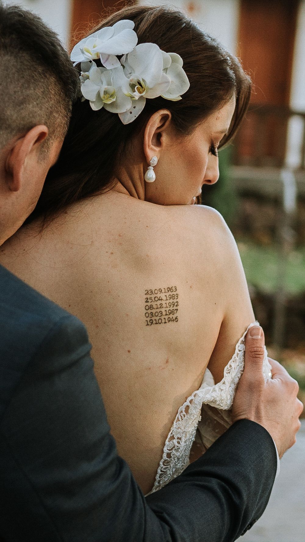 Groom embraces bride from behind revealing meaningful date tattoo on her bare back, adorned with orchid hair flowers.