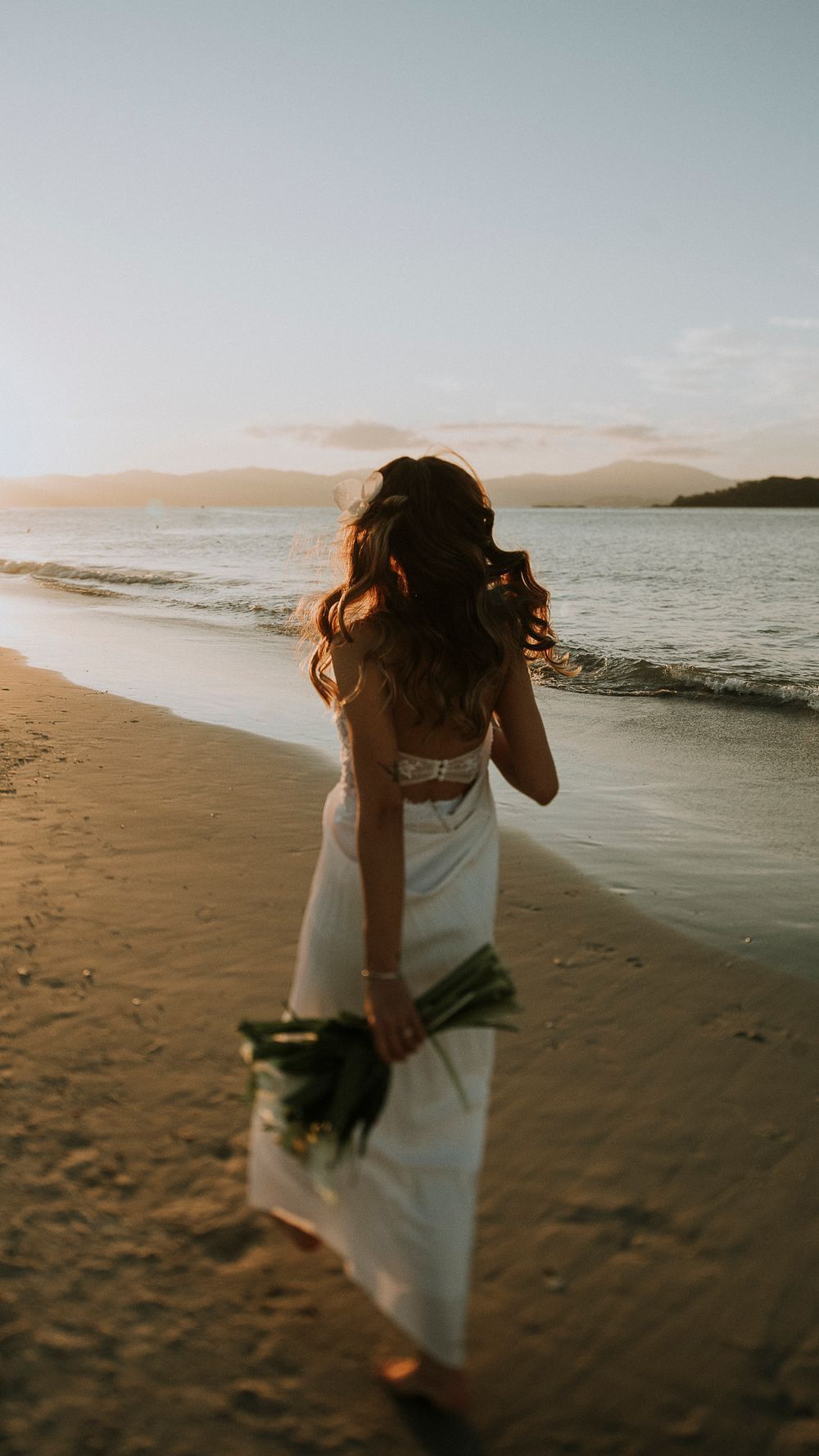 Bride in white gown holding a bouquet walks along a golden sunset beach shoreline.