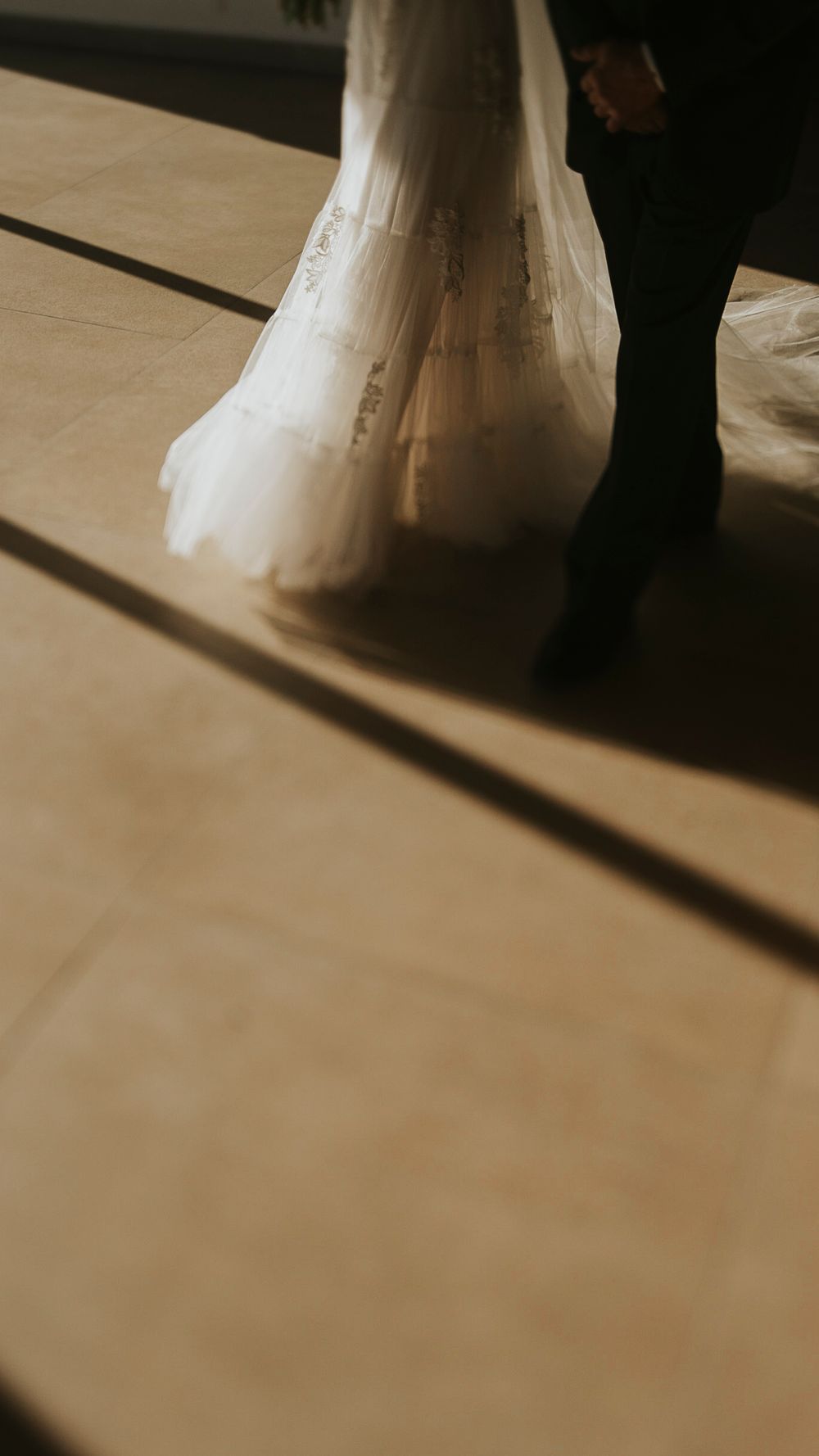Close-up of bride in white lace wedding gown and groom in dark suit standing on sunlit tiled floor.