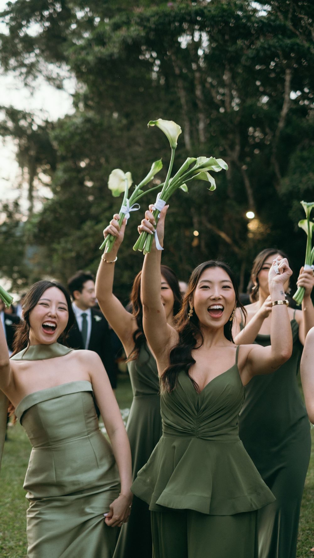 Excited bridesmaids in mismatched olive green gowns raise calla lily bouquets in celebration at outdoor wedding.