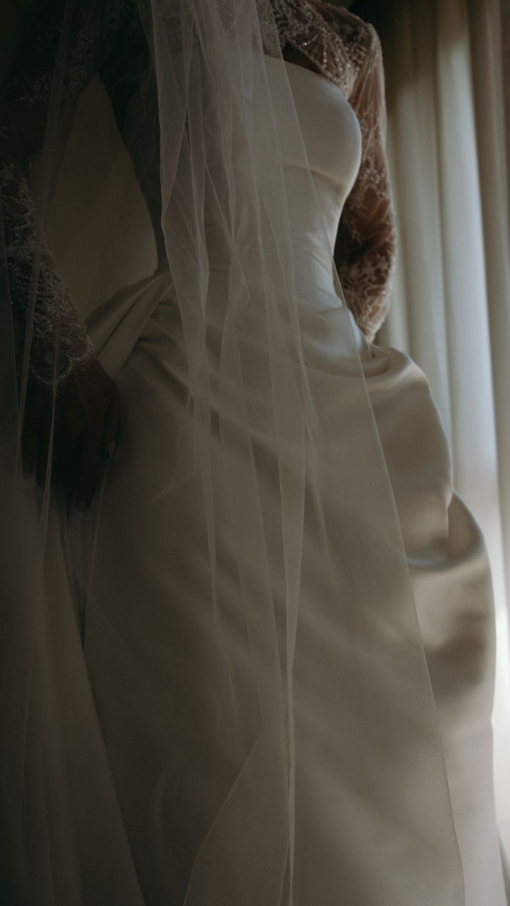 Close-up of a white satin wedding gown with lace veil draped by a soft curtained window.