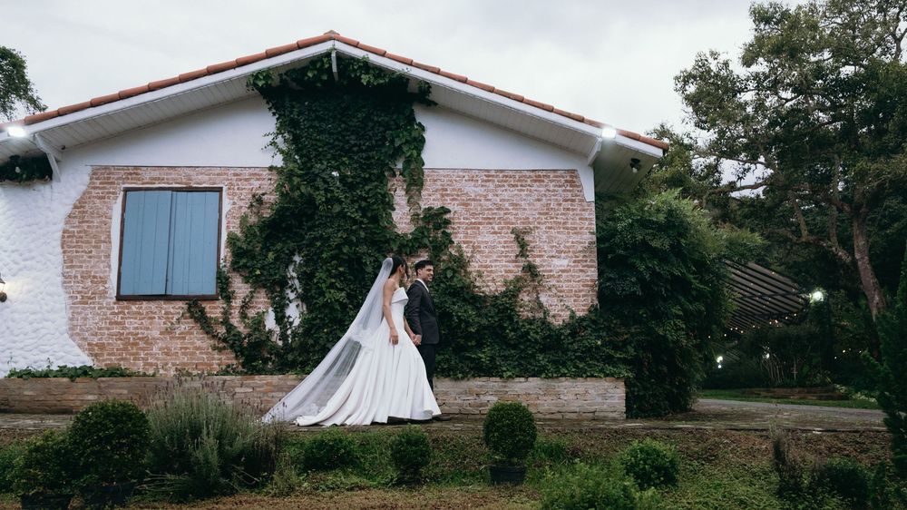 Bride in flowing white gown with long veil standing before ivy-covered brick house outdoors.