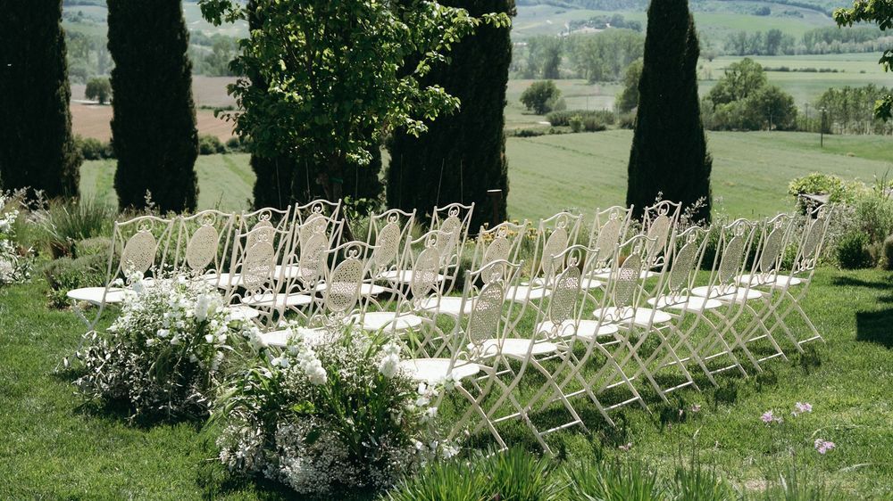 Rows of white folding chairs set outdoors among cypress trees for elegant garden wedding ceremony.